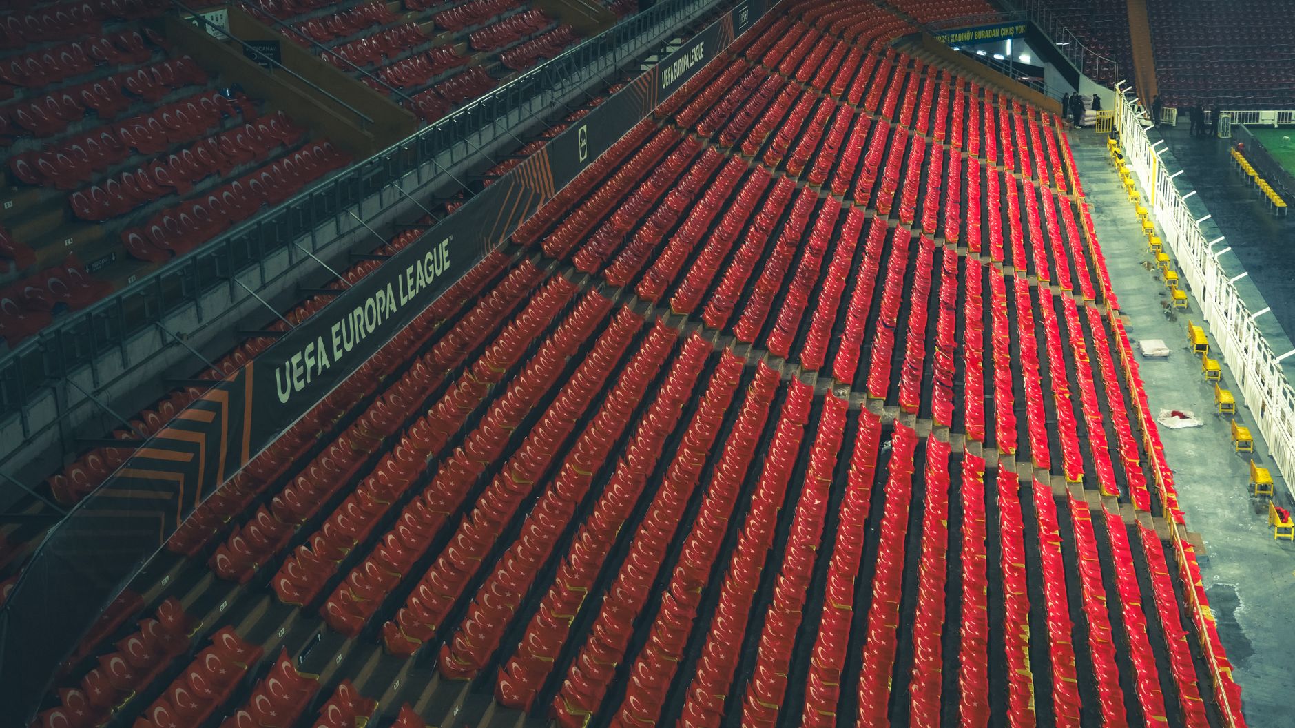 rows of empty red stadium seats inside a large football arena, floodlights illuminating the green pitch below