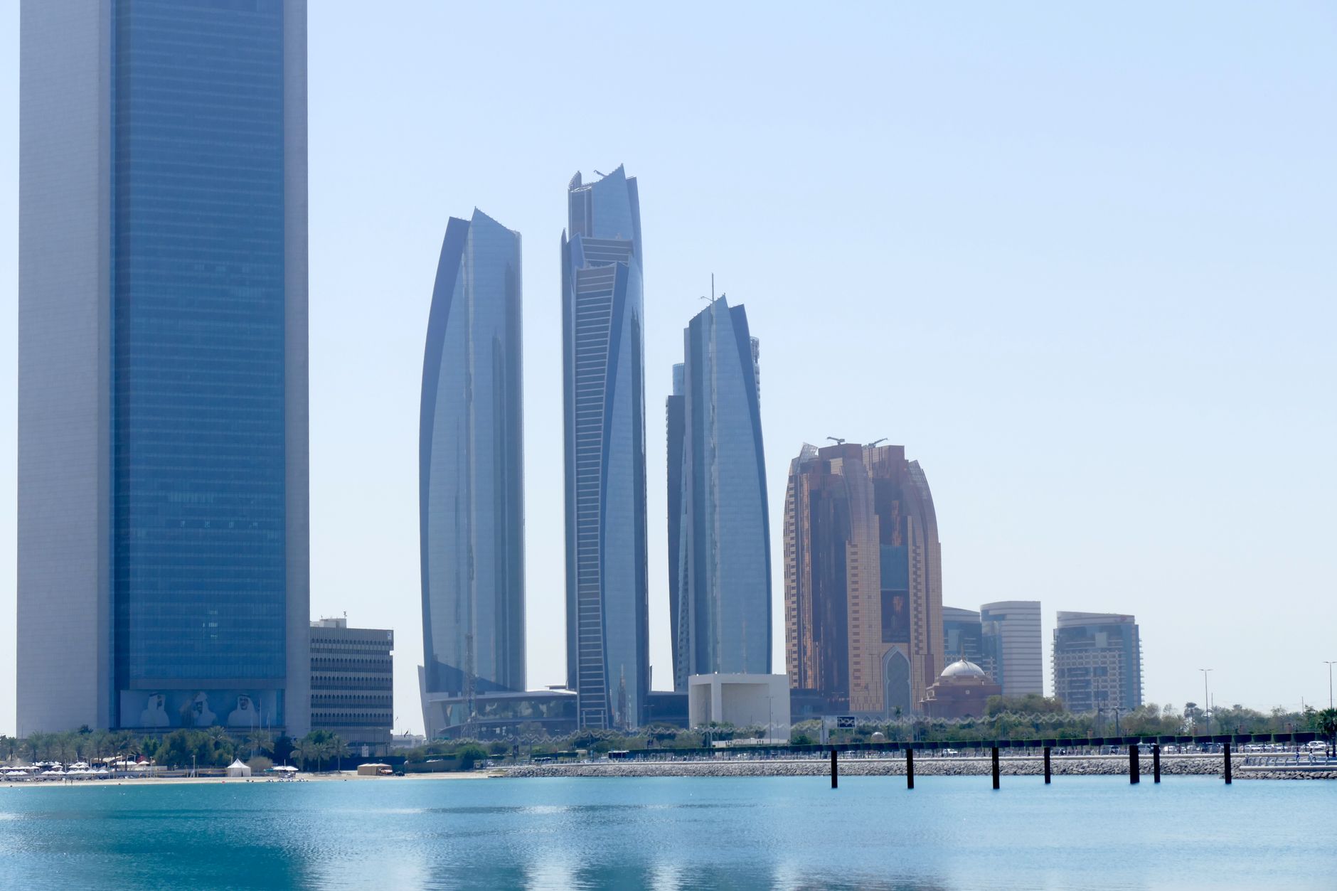 wide shot of a major Gulf financial district at midday showing gleaming institutional towers reflected in a calm waterway, with construction activity visible in the background