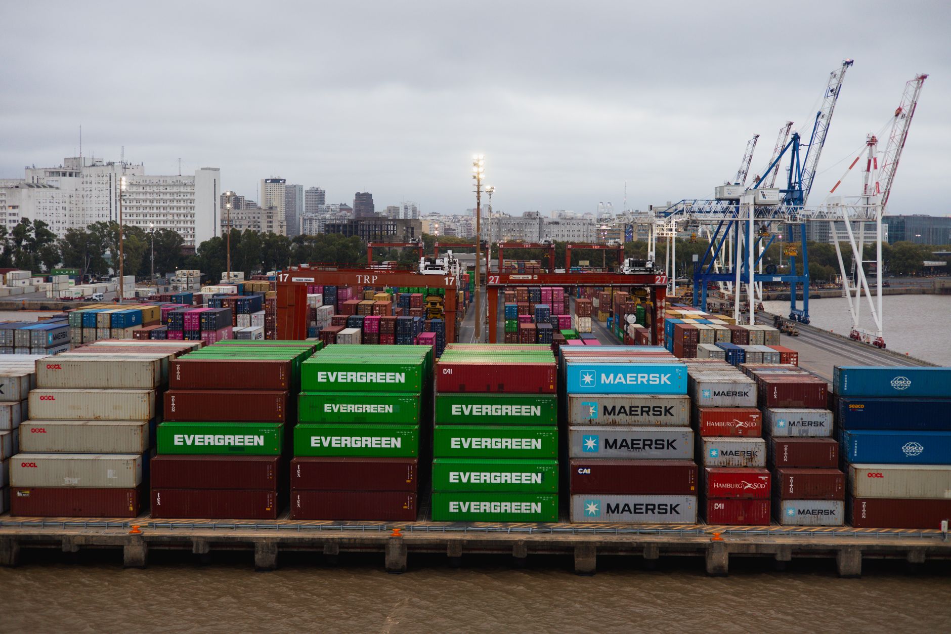 rows of shipping containers stacked at a busy South Korean port terminal, with large cargo cranes visible against a grey overcast sky