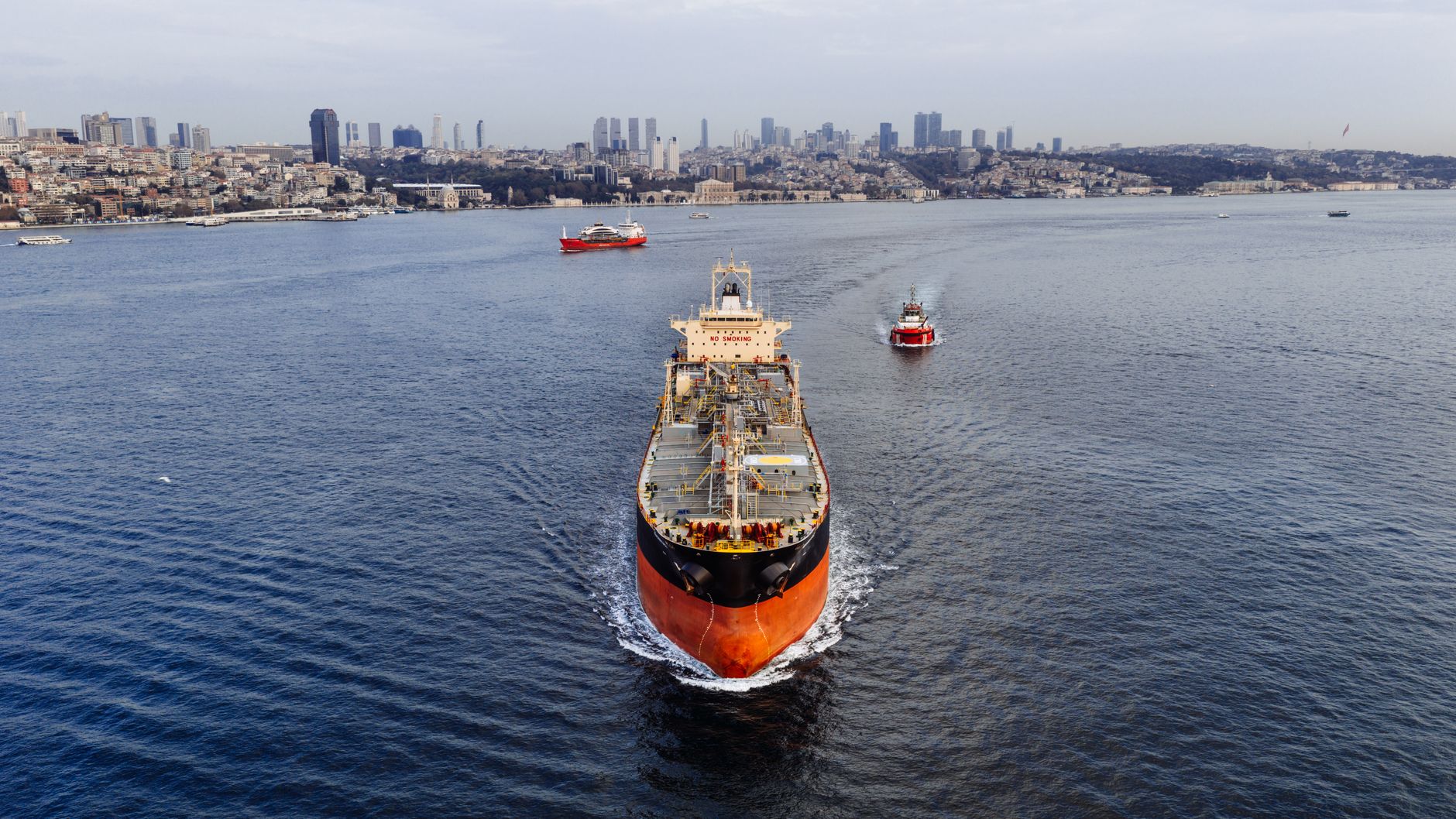 A large oil tanker anchored in still open water near a narrow strait, viewed from above at distance with a coastline visible on the horizon
