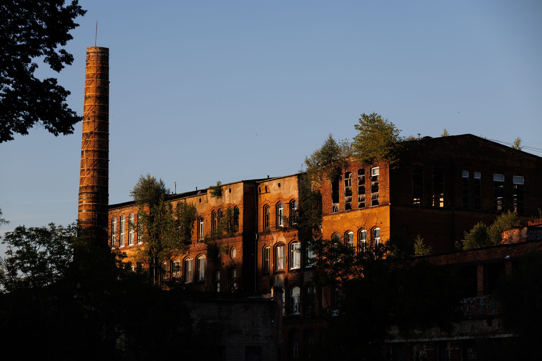 exterior of a Victorian-era red-brick factory building with tall chimneys, set against an overcast sky, surrounded by overgrown cobblestones