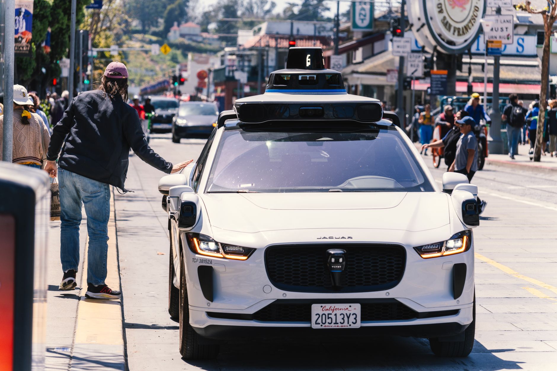 a Waymo robotaxi vehicle parked at a curbside charging station on a quiet urban street at dusk, no occupants visible, city buildings reflected in the vehicle's windows