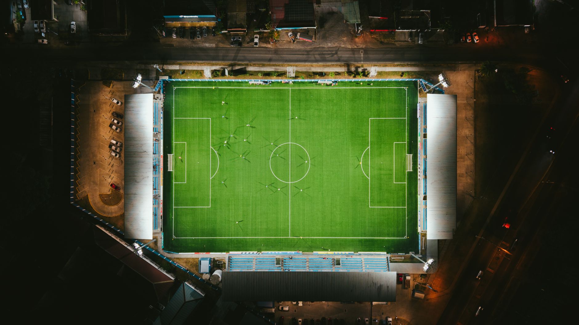wide aerial view of a large college football stadium at dusk, with the field illuminated under stadium lights and surrounding campus buildings visible in the background