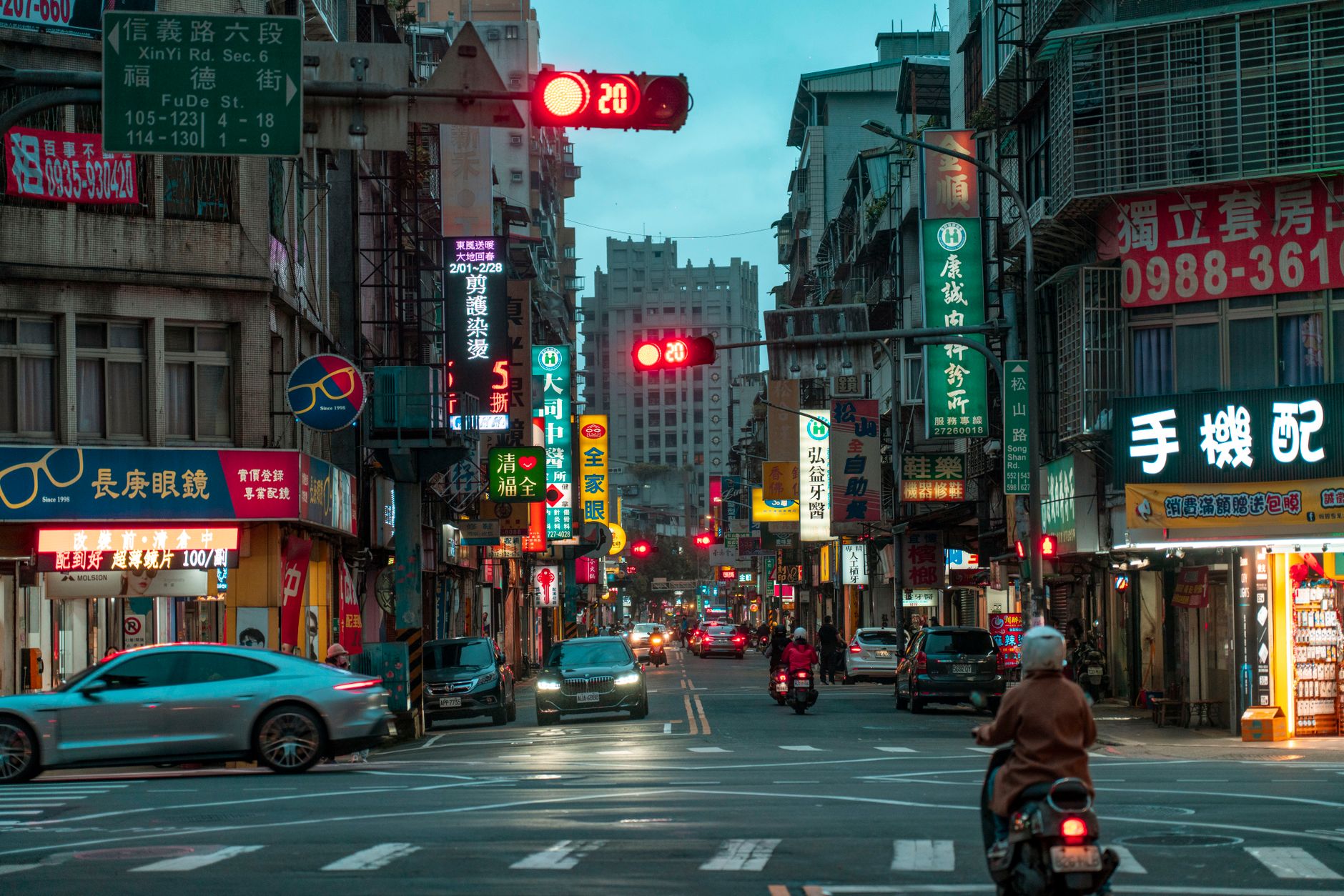 a busy commercial street in a Chinese city at dusk, storefronts illuminated, with delivery vehicles and pedestrians visible in the background as small incidental figures