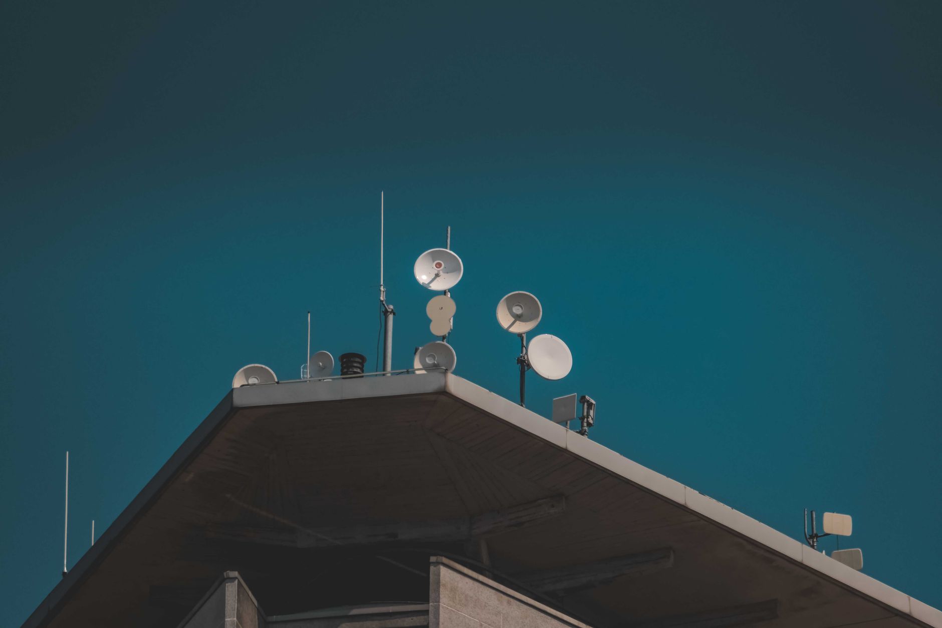 exterior of a modern defense systems integration facility at dusk, with satellite dishes and antenna structures silhouetted against an orange sky