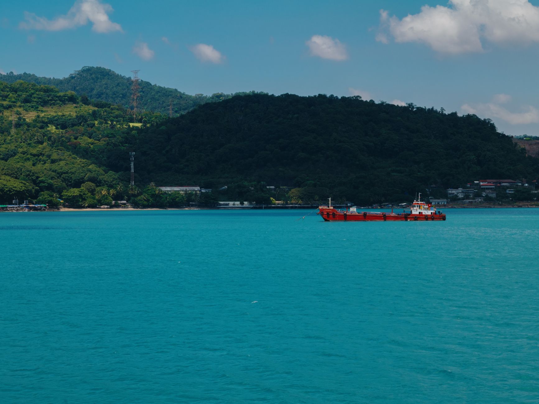 a commercial oil tanker at anchor in calm blue waters near a rocky coastline, with cargo cranes visible on the distant shore