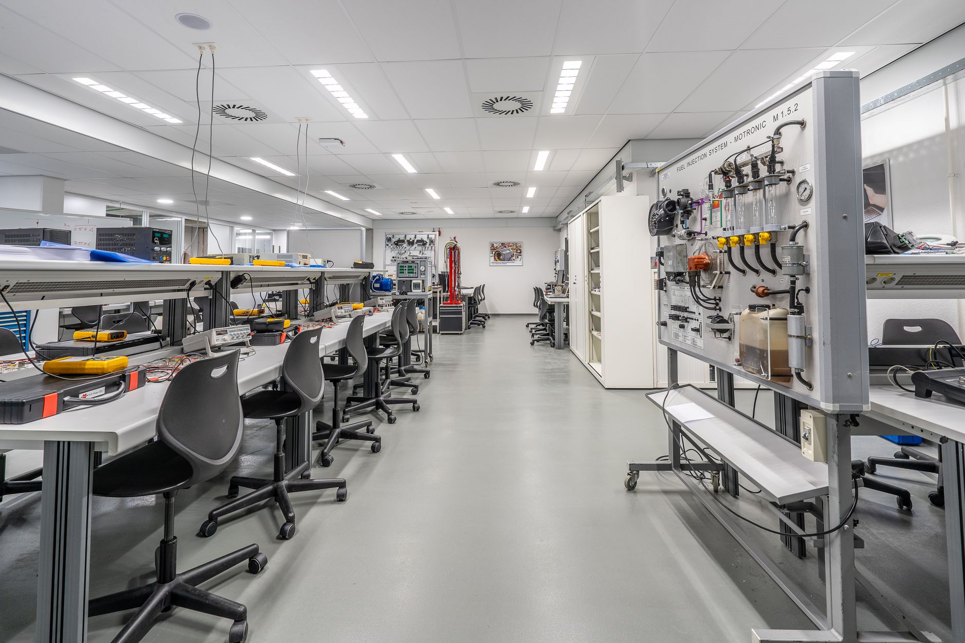 rows of modular battery cell testing equipment inside a clean-room industrial laboratory, with white flooring and overhead fluorescent lighting reflecting off metallic surfaces