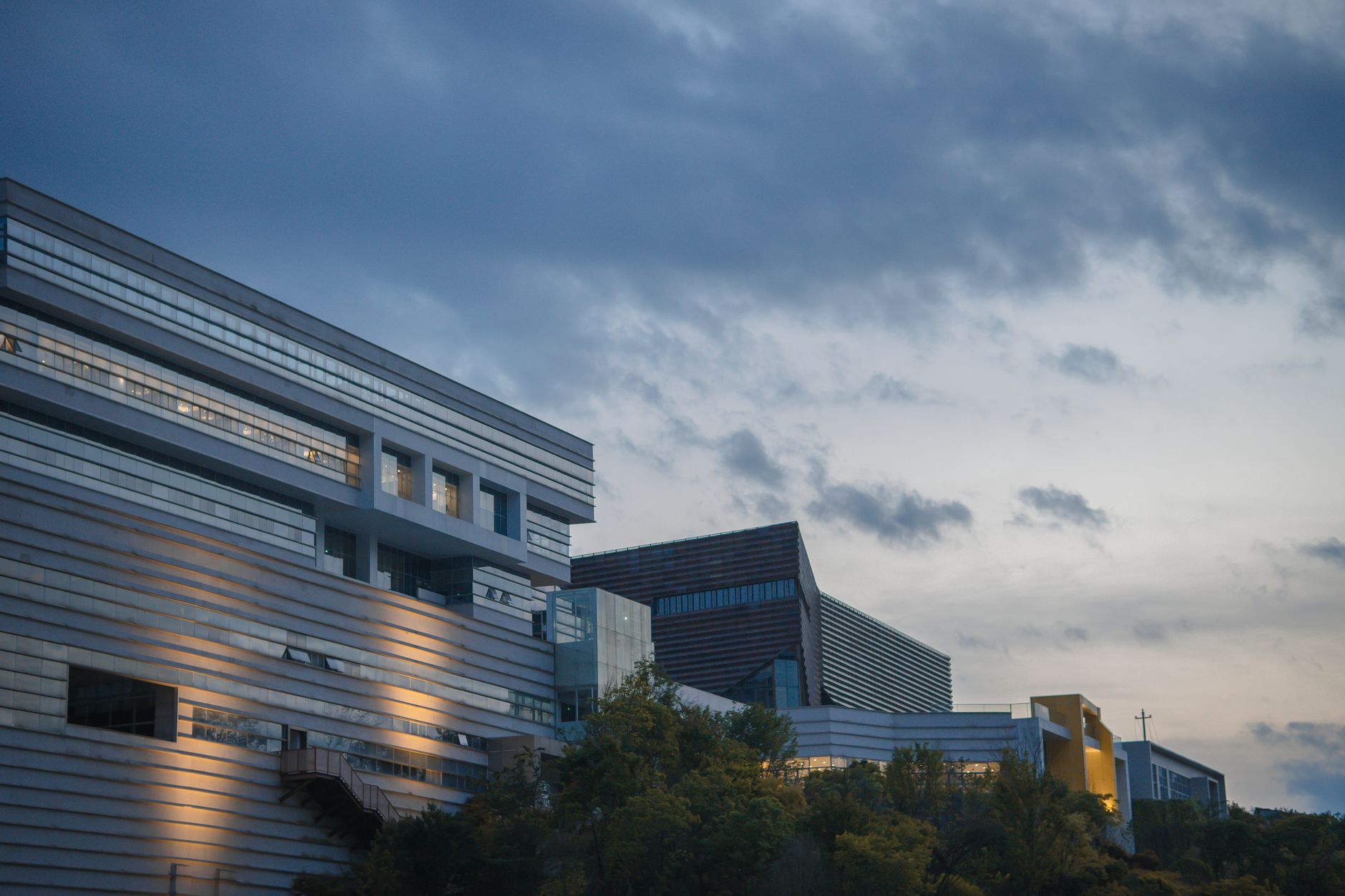 exterior of a modern European research campus at dusk, featuring low-profile glass and steel buildings surrounded by manicured grounds, with illuminated windows casting warm light against a darkening sky
