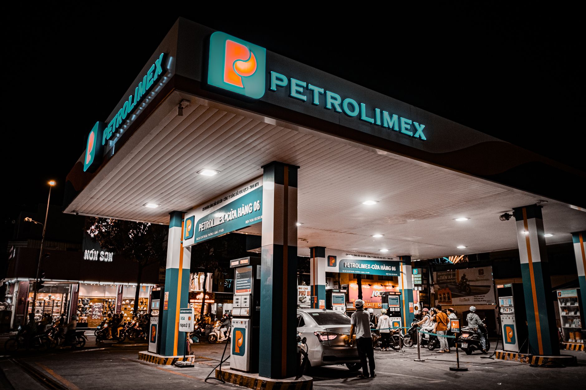 exterior of a busy Tehran petrol station forecourt with rows of fuel pumps and queuing vehicles under fluorescent canopy lighting at dusk