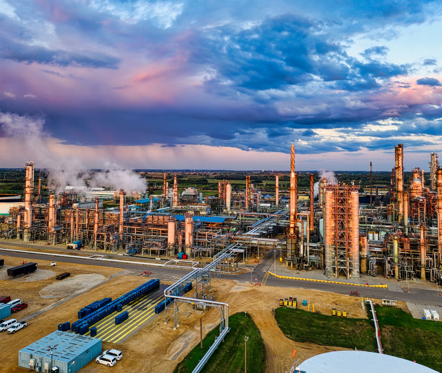 exterior of a large petrochemical refinery at dusk with smokestacks and storage tanks visible against an orange sky
