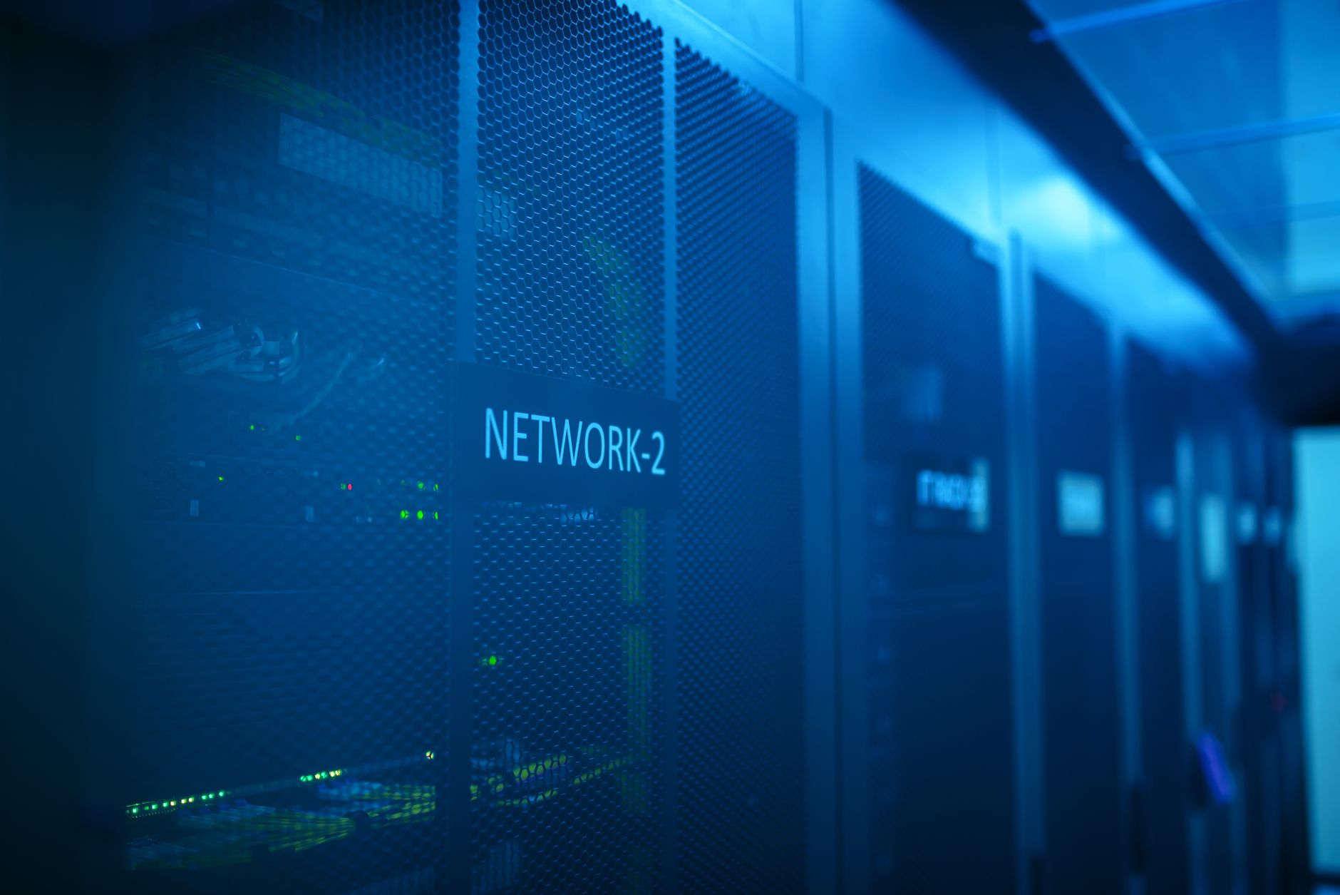 rows of illuminated server racks inside a large data center facility, viewed from a low angle with cooling infrastructure visible overhead