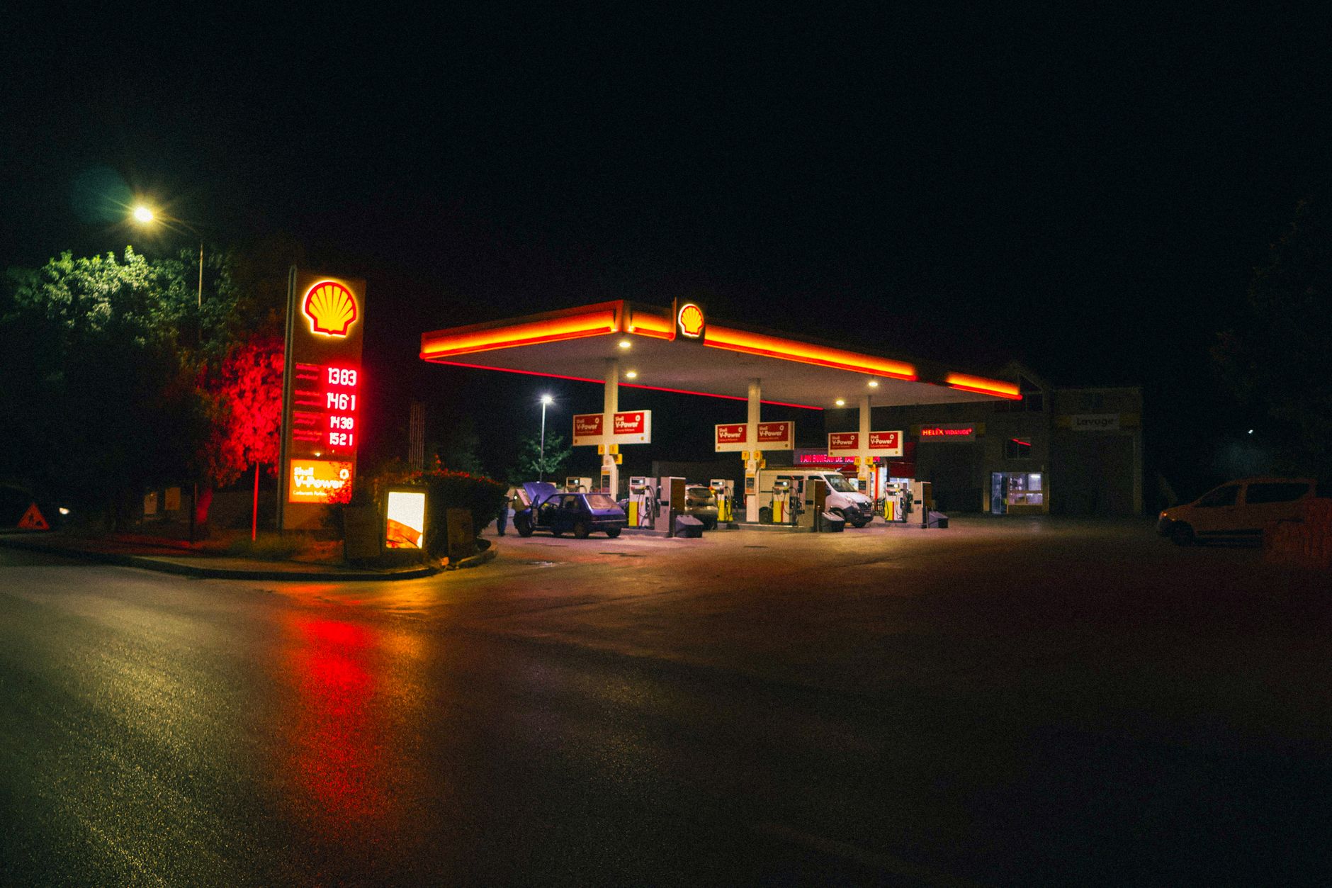 exterior of a modern fuel station forecourt at night with illuminated signage and a convenience store entrance visible through glass doors