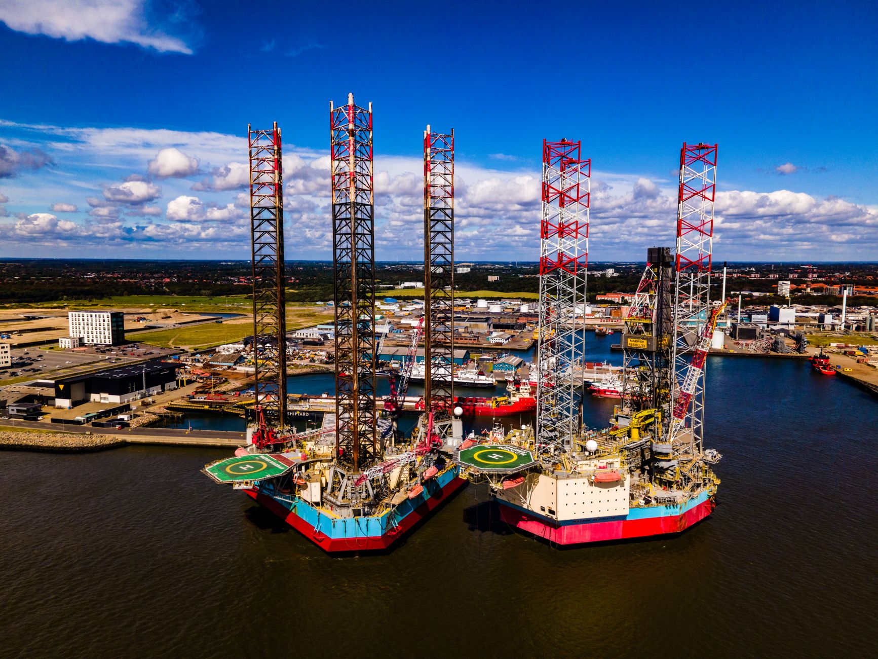 wide aerial view of a damaged industrial port facility along a coastline, cranes tilted and infrastructure visibly disrupted, no people in foreground