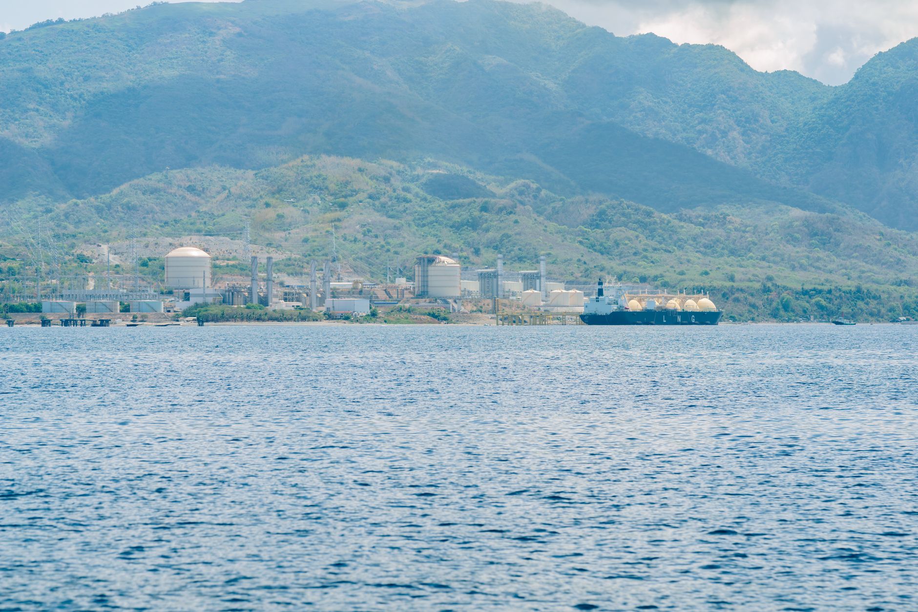 a cargo tanker navigating a narrow strait at low tide, surrounded by open sea with overcast skies and no visible port infrastructure