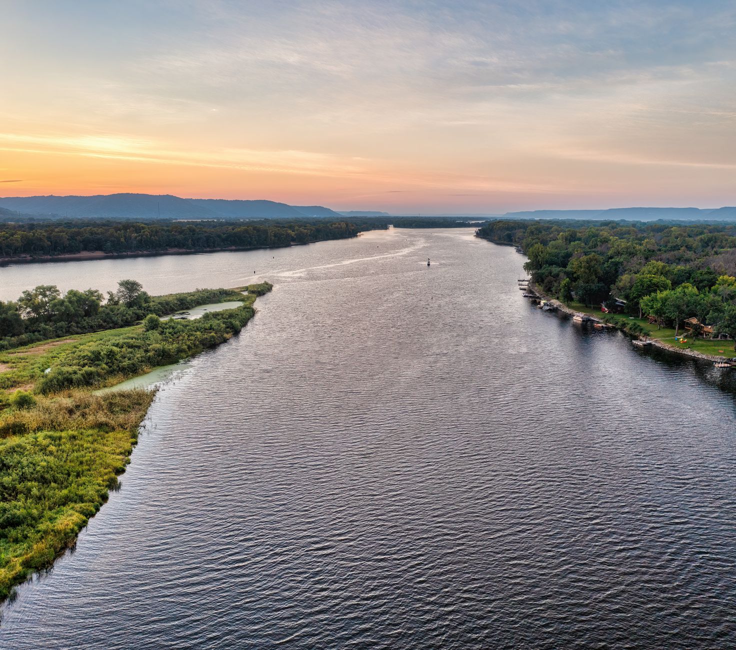 aerial view of a large North American lake at dawn, calm water reflecting early morning sky with forested shoreline in the background