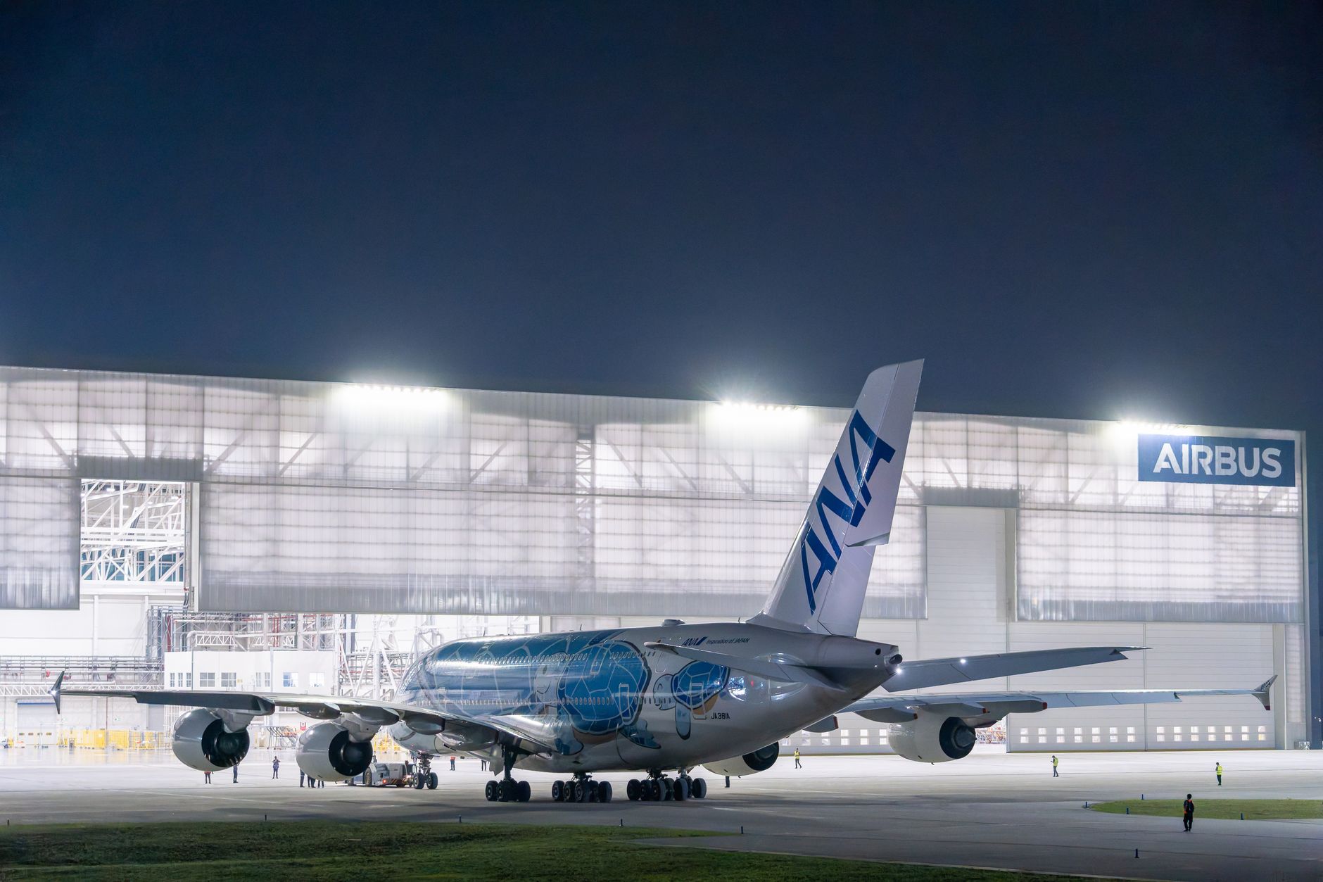 exterior of a mid-sized aerospace manufacturing plant in an industrial zone, with aircraft ground support vehicles visible on the tarmac in the background