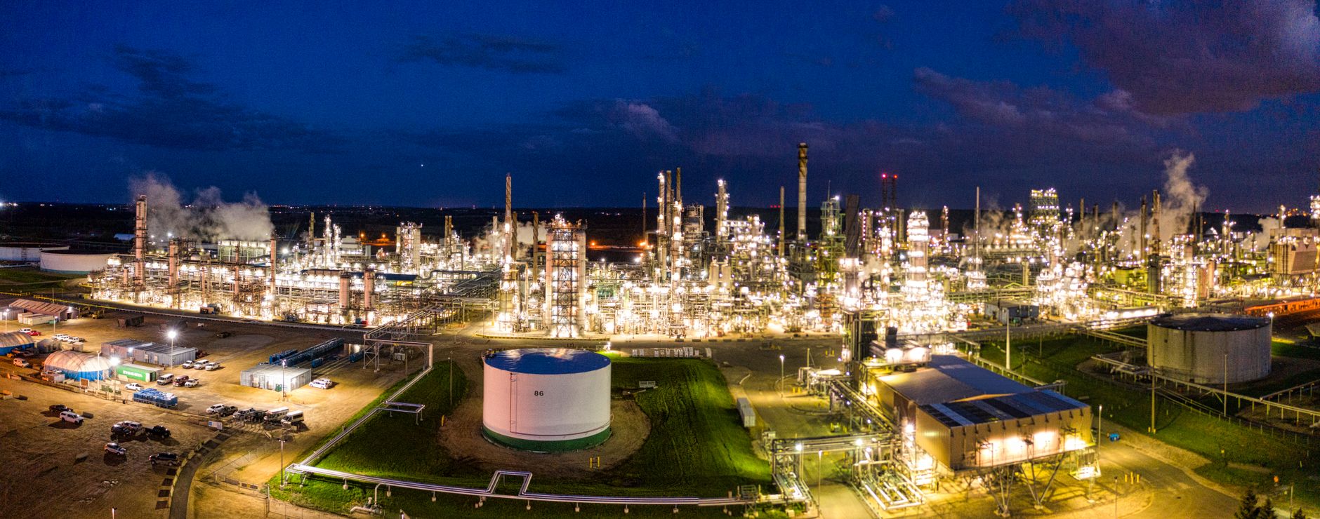aerial view of a sprawling Middle Eastern oil refinery complex at dusk, with lit processing towers and pipeline infrastructure visible across a flat desert landscape