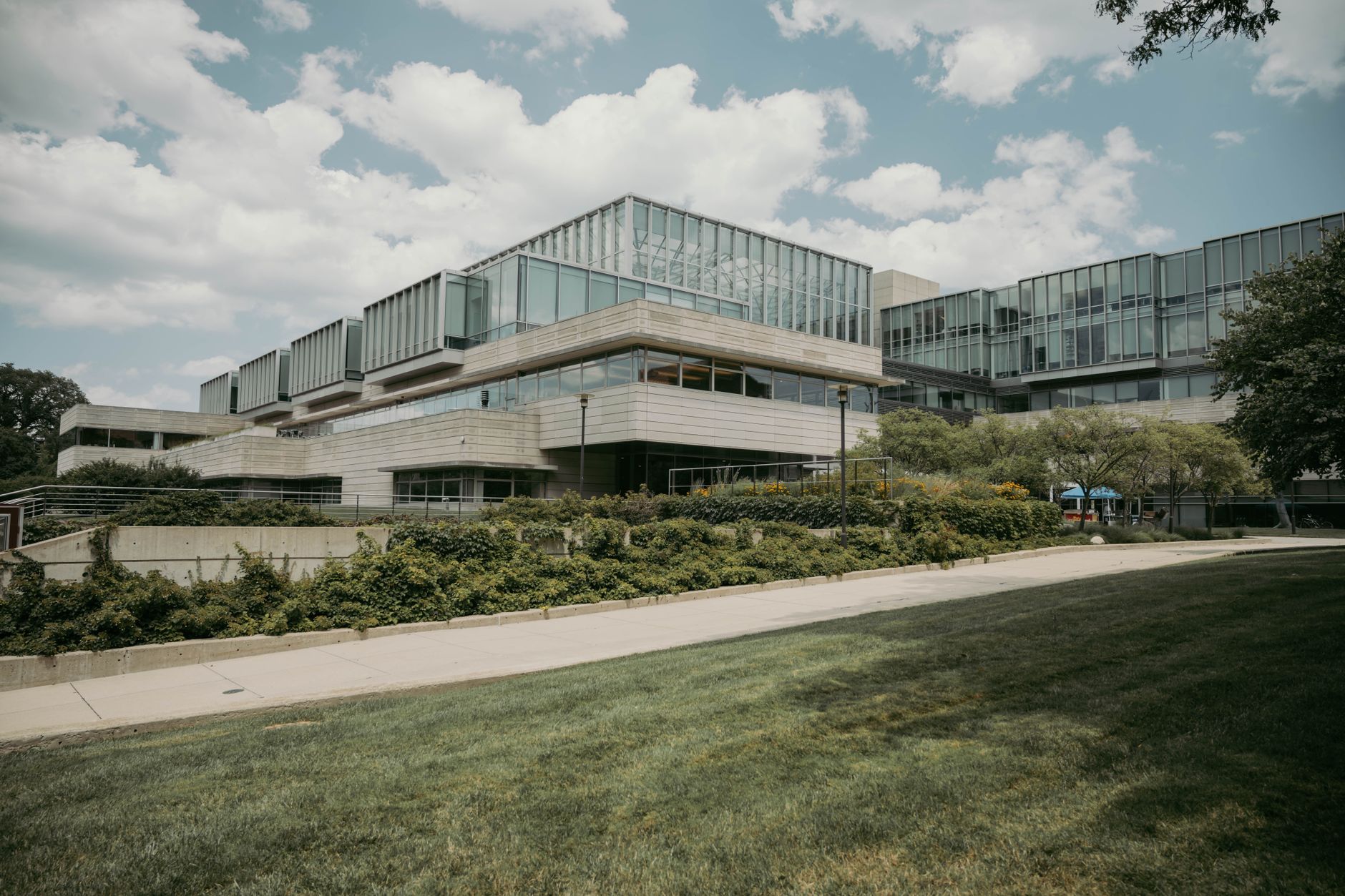 exterior view of a pharmaceutical research campus at dusk, glass-and-steel buildings surrounded by manicured grounds, cargo trucks visible at a loading bay in the background