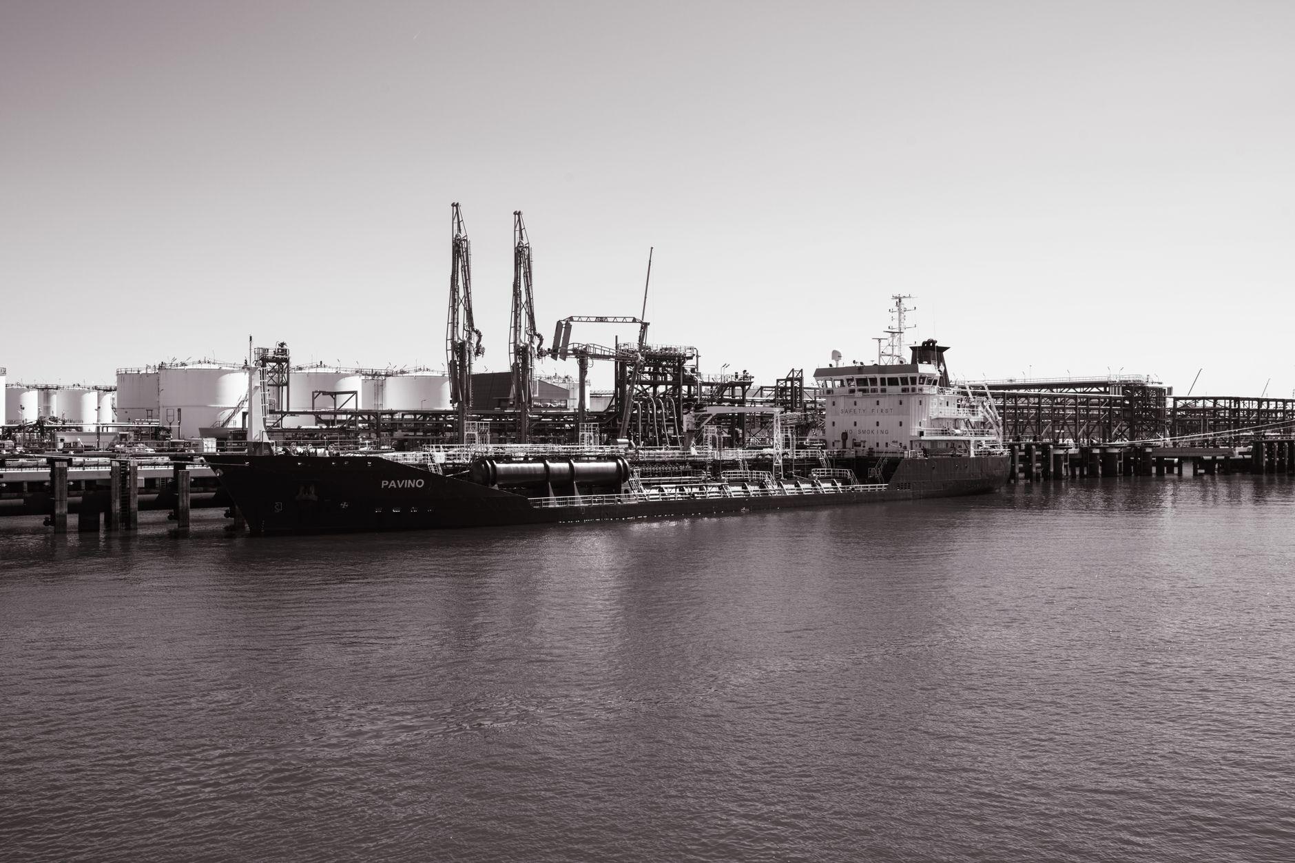 a large oil tanker anchored in a gray industrial port, visible refinery infrastructure and storage tanks along the waterfront in the distance