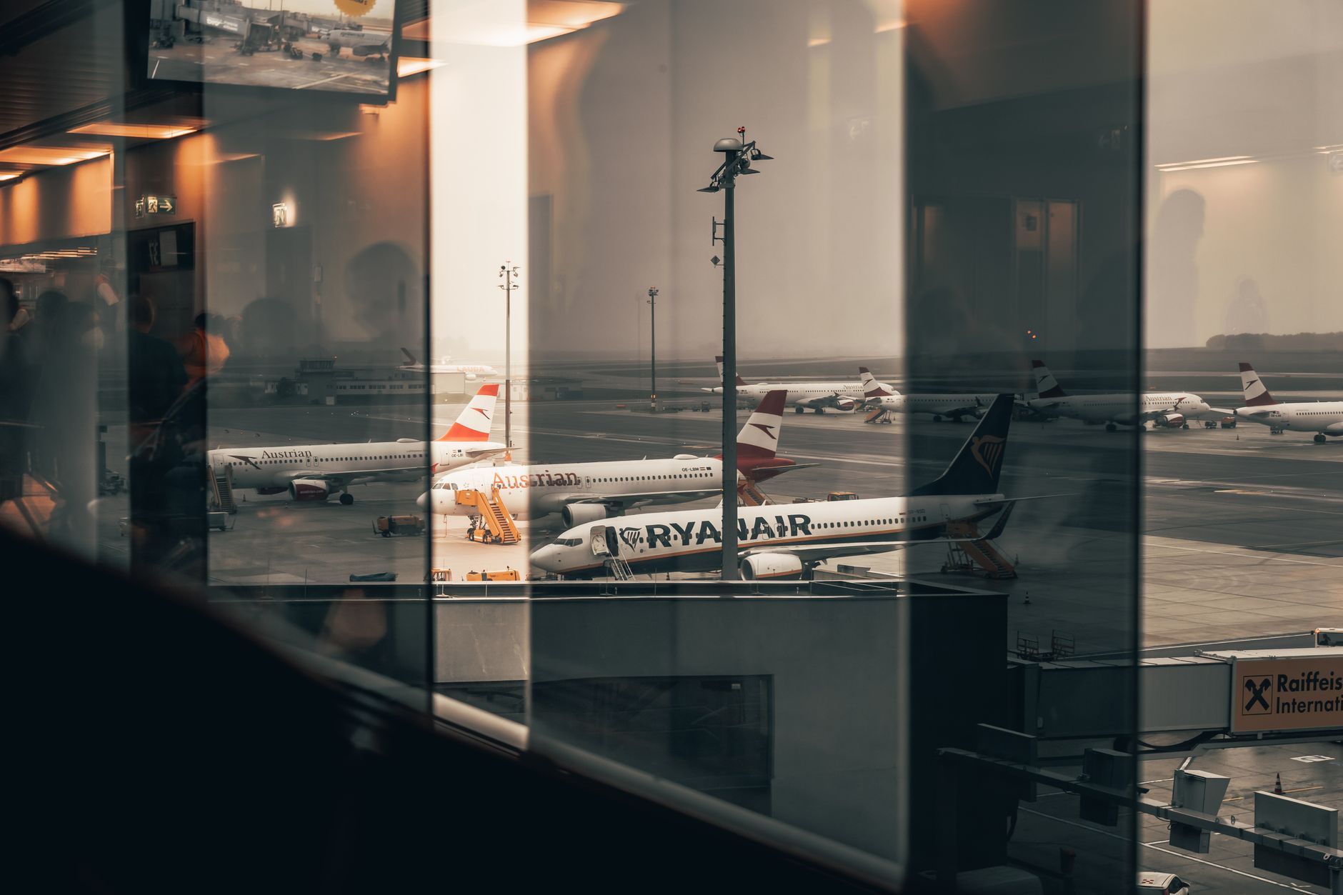 wide-angle view of a busy international airport terminal at dusk, with large aircraft visible through floor-to-ceiling glass windows and departure boards displaying multiple global city names