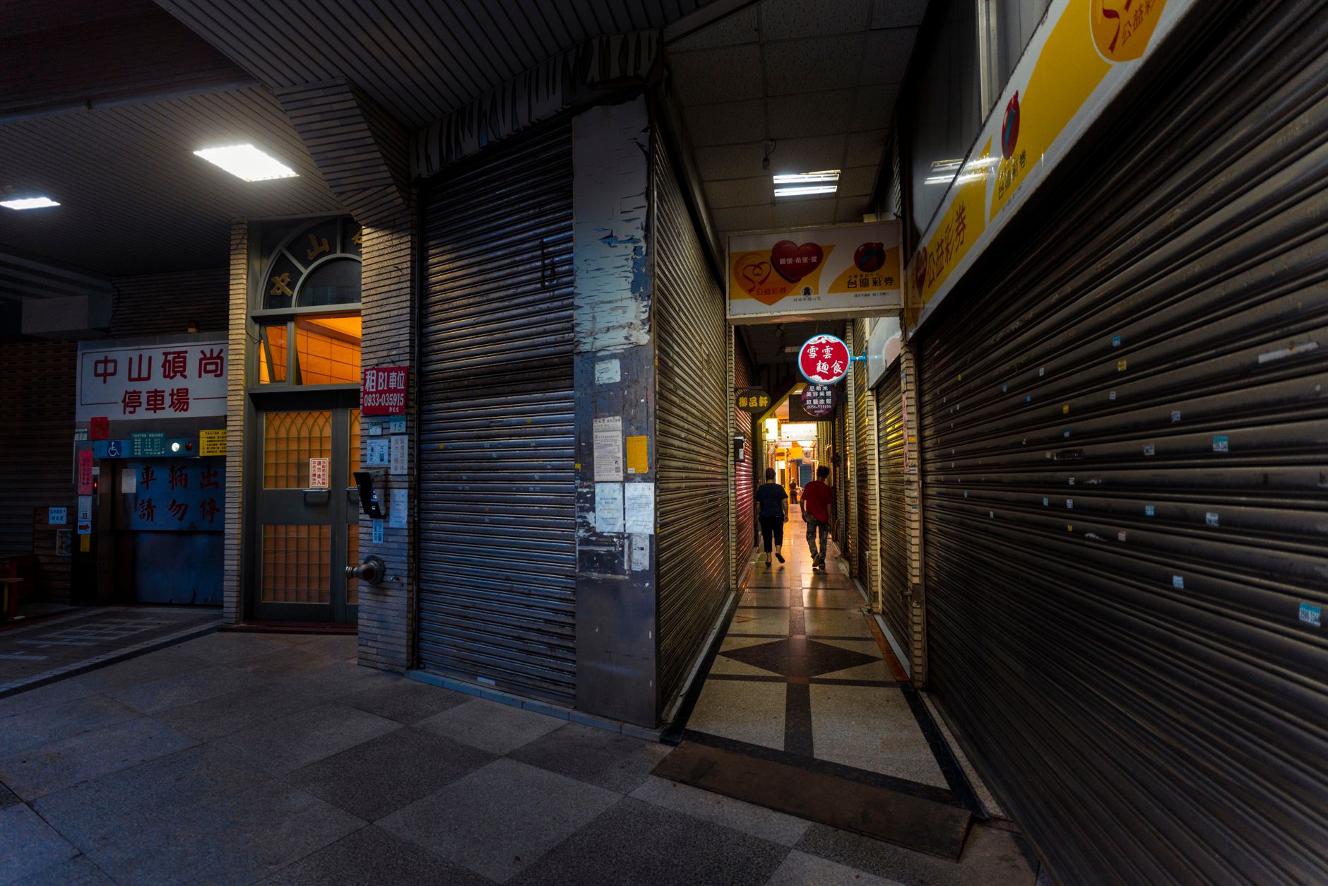 rows of closed metal shutters along a commercial street in Beirut, faded signage visible on storefronts, afternoon light casting long shadows