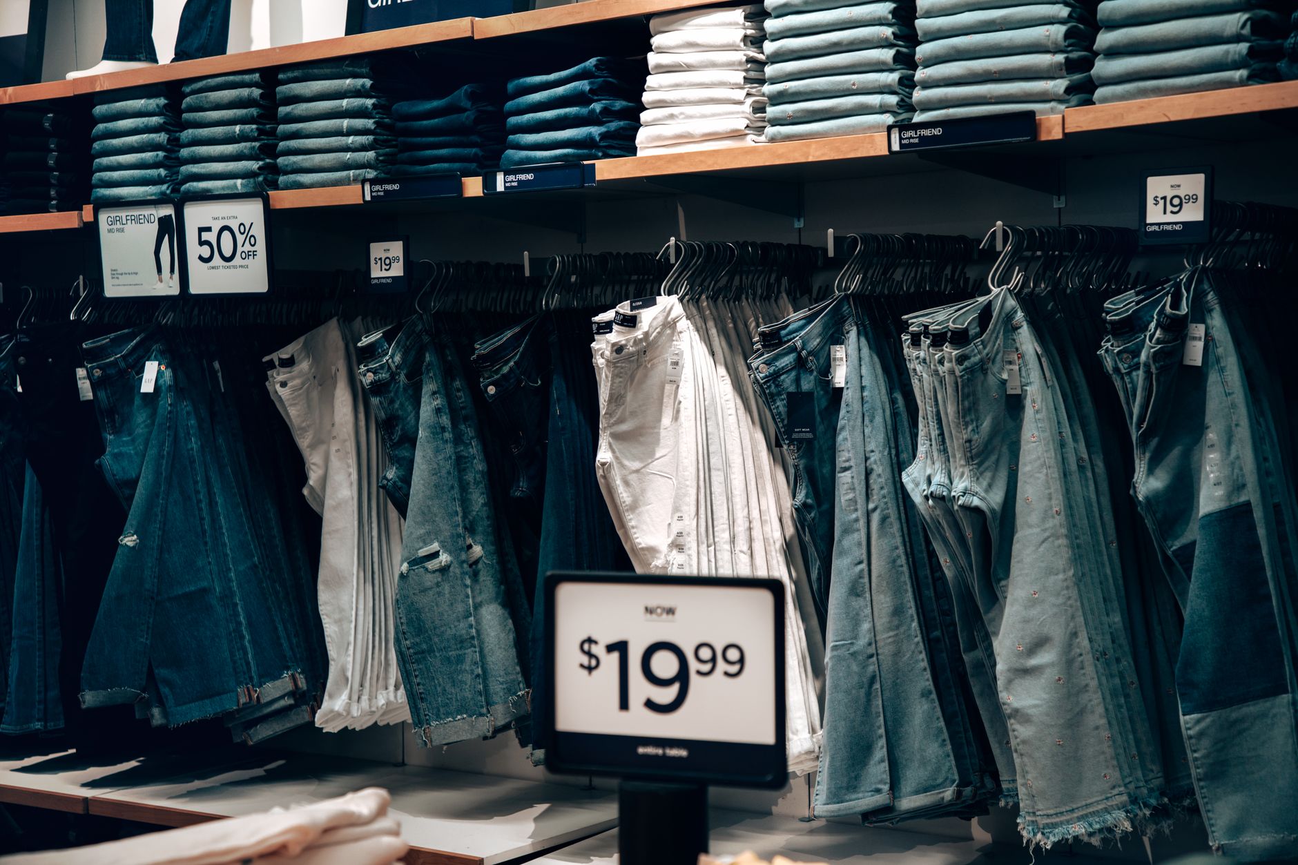 rows of retail clothing racks in a large department store, with sale signs on display and sparse customer activity visible in the background