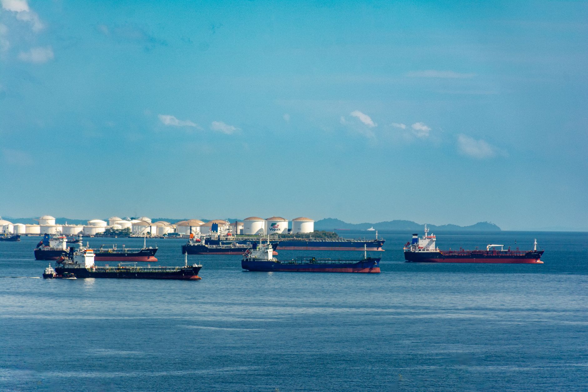 aerial view of oil tankers anchored in formation near a narrow strait, shot from a wide altitude with blue-green water and distant coastline visible