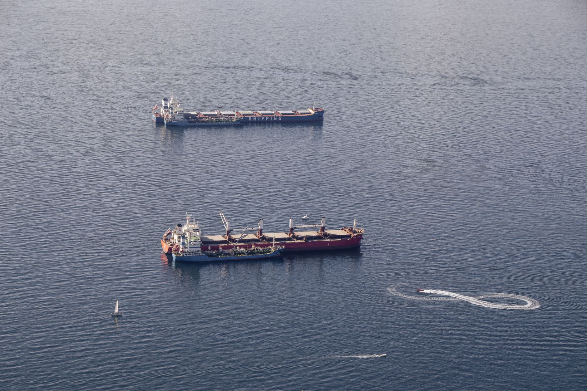aerial view of an oil tanker navigating a narrow strait, surrounded by calm blue water under a hazy sky