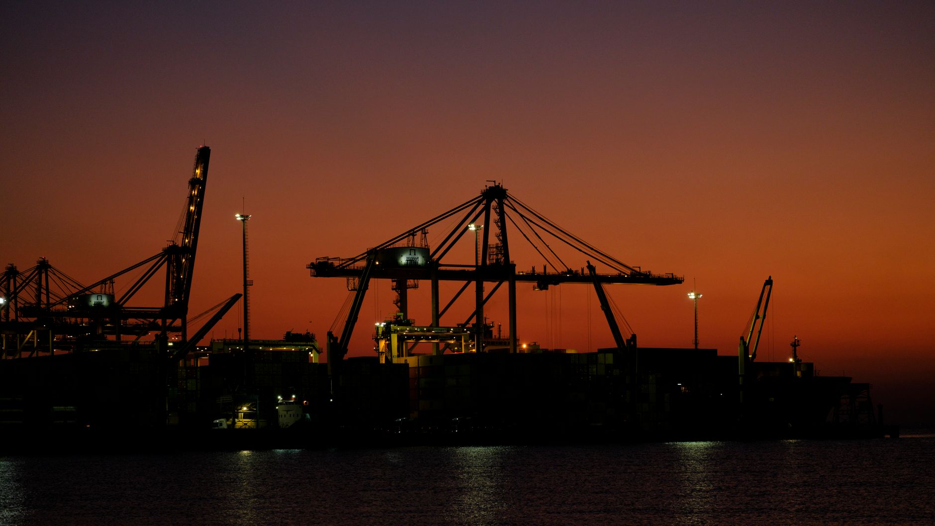 exterior of a large port facility at dusk, with rows of shipping containers and cranes silhouetted against an orange sky