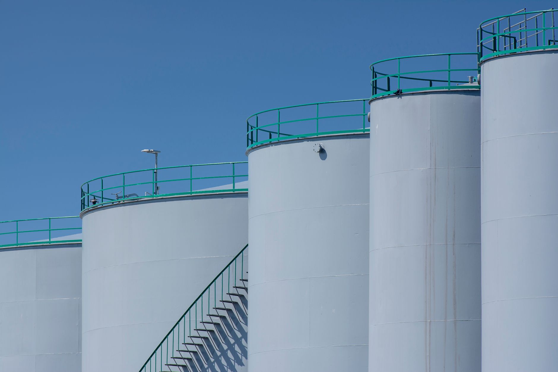 rows of oil storage tanks at a coastal petroleum terminal, viewed from an elevated angle at dusk with tanker vessels visible in the distance