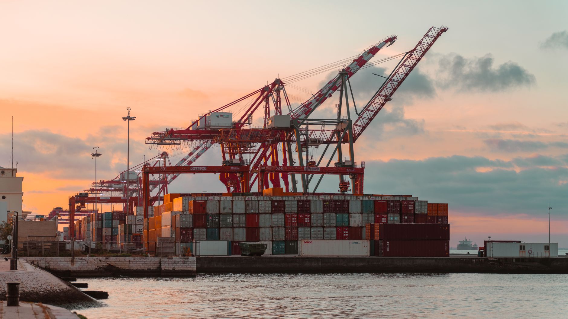 container ships moored at a busy Vietnamese industrial port at dusk, with cargo cranes silhouetted against an orange sky