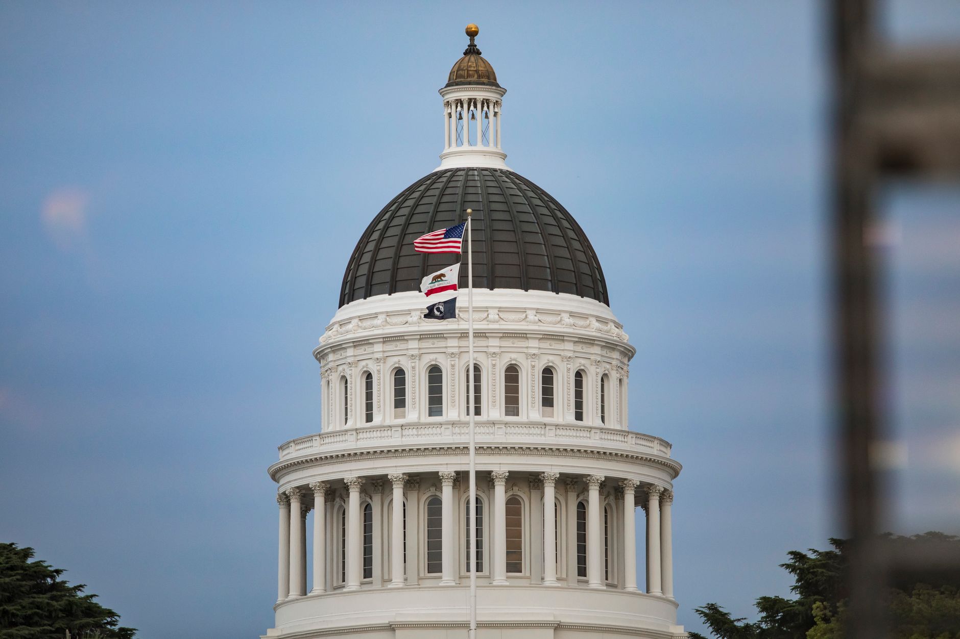 exterior of a state capitol building at midday with an empty plaza and flagpoles in the foreground, wide-angle perspective