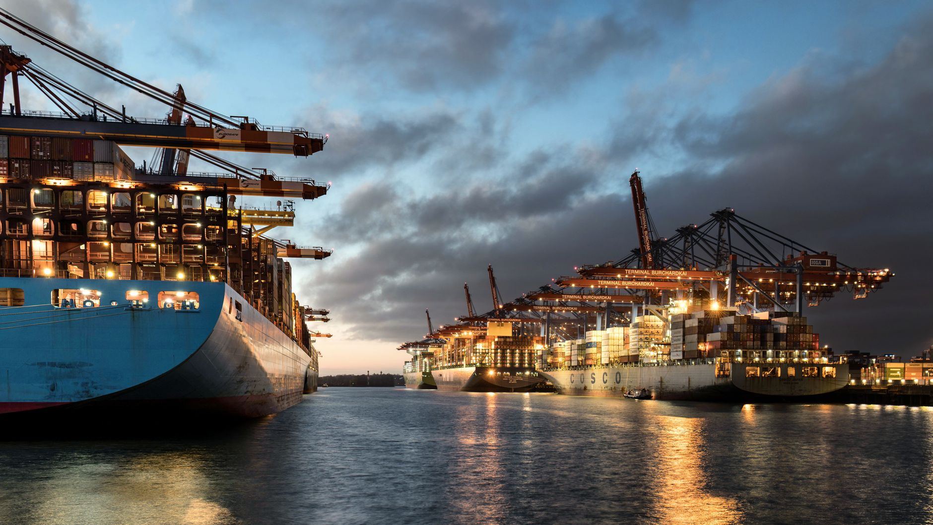wide-angle view of the Port of Alexandria at dusk, showing large container ships docked at industrial berths with cranes overhead