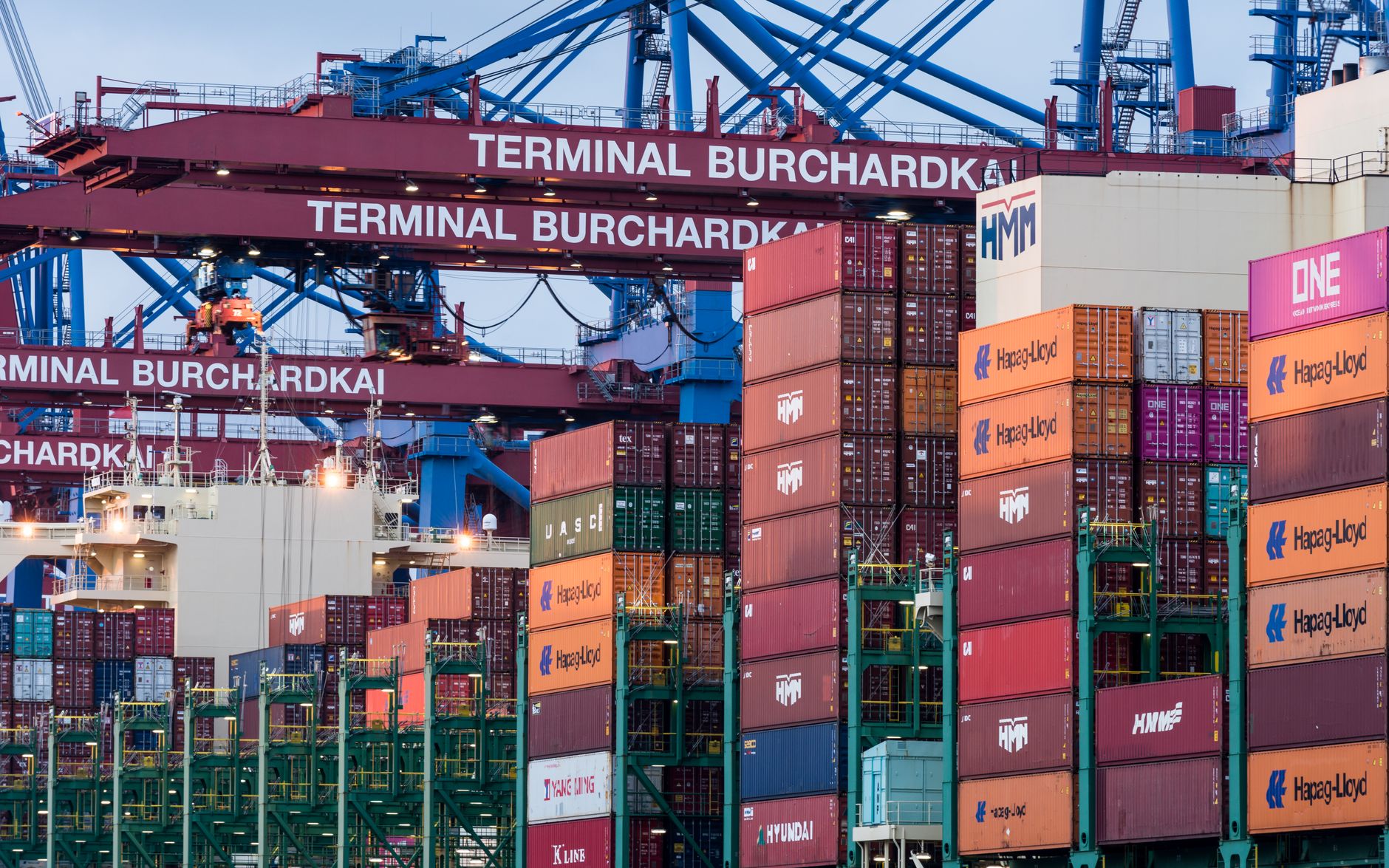 rows of shipping containers stacked at a large coastal port terminal, cargo cranes silhouetted against an overcast sky