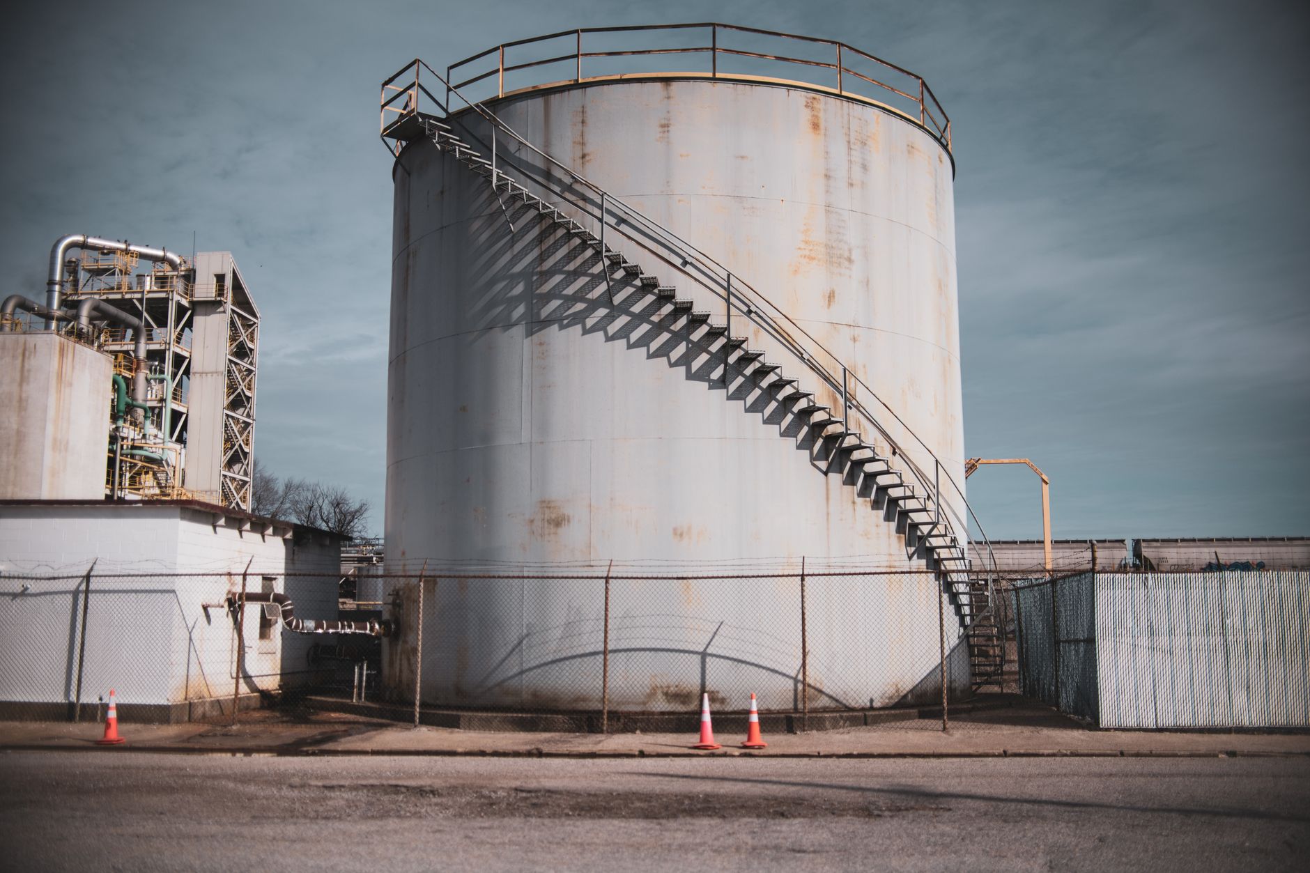 rows of oil storage tanks in an industrial refinery complex photographed from above at dusk, pipes and infrastructure stretching to the horizon