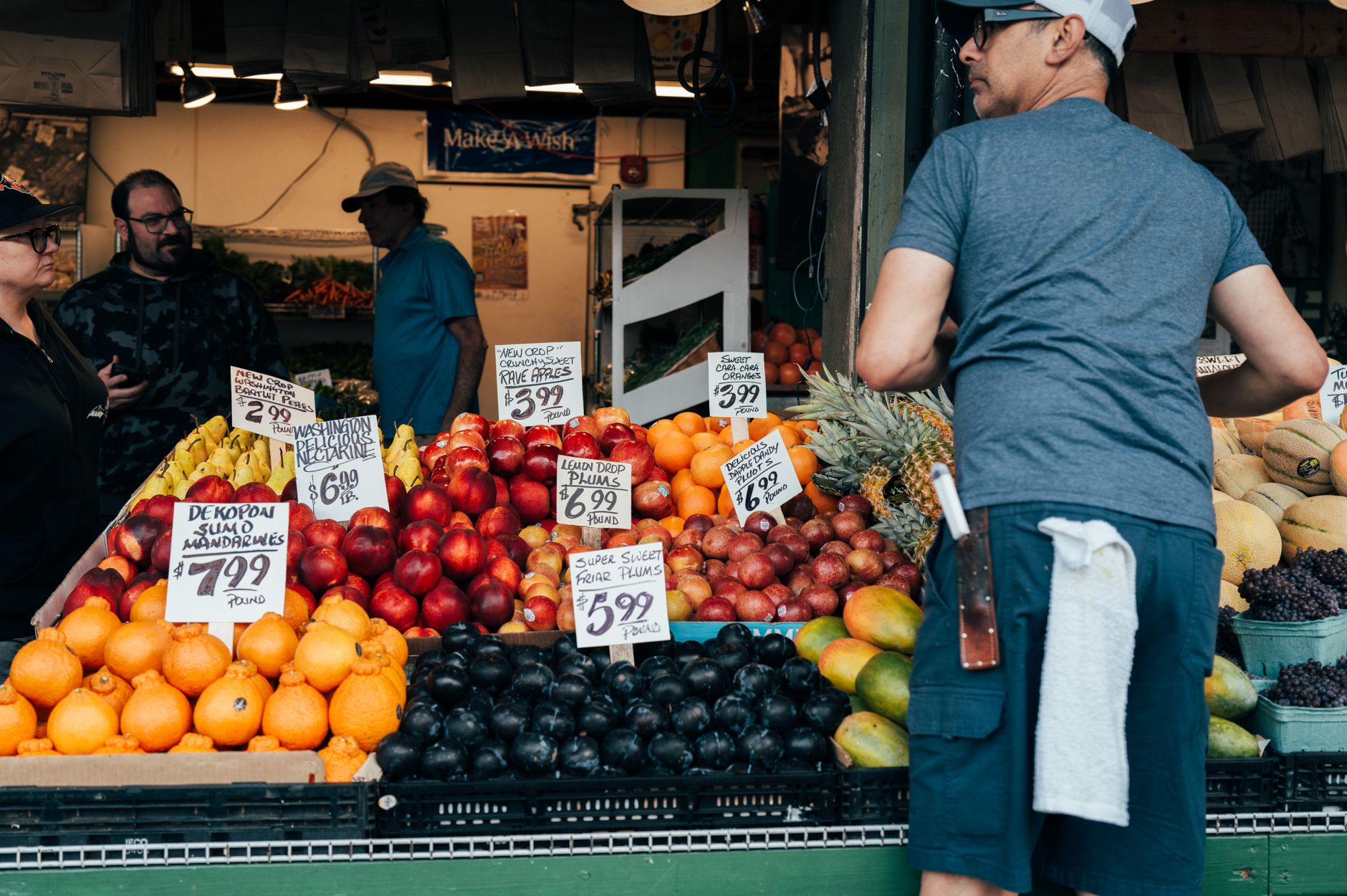 a busy outdoor vegetable market with crates of produce and handwritten price boards visible on stall fronts