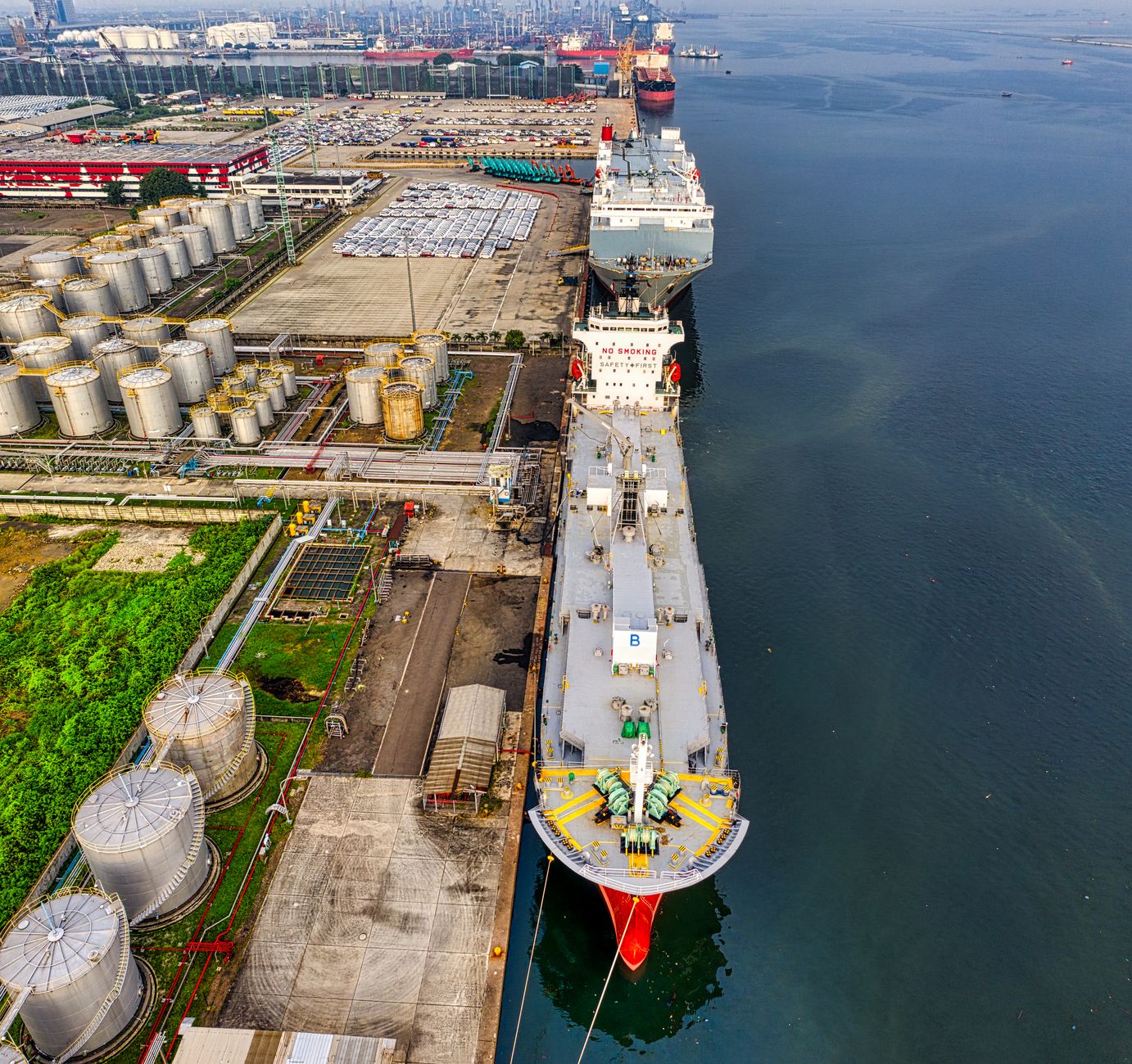 aerial view of large oil tankers navigating through a narrow sea strait at dusk, surrounded by industrial port infrastructure on both shorelines