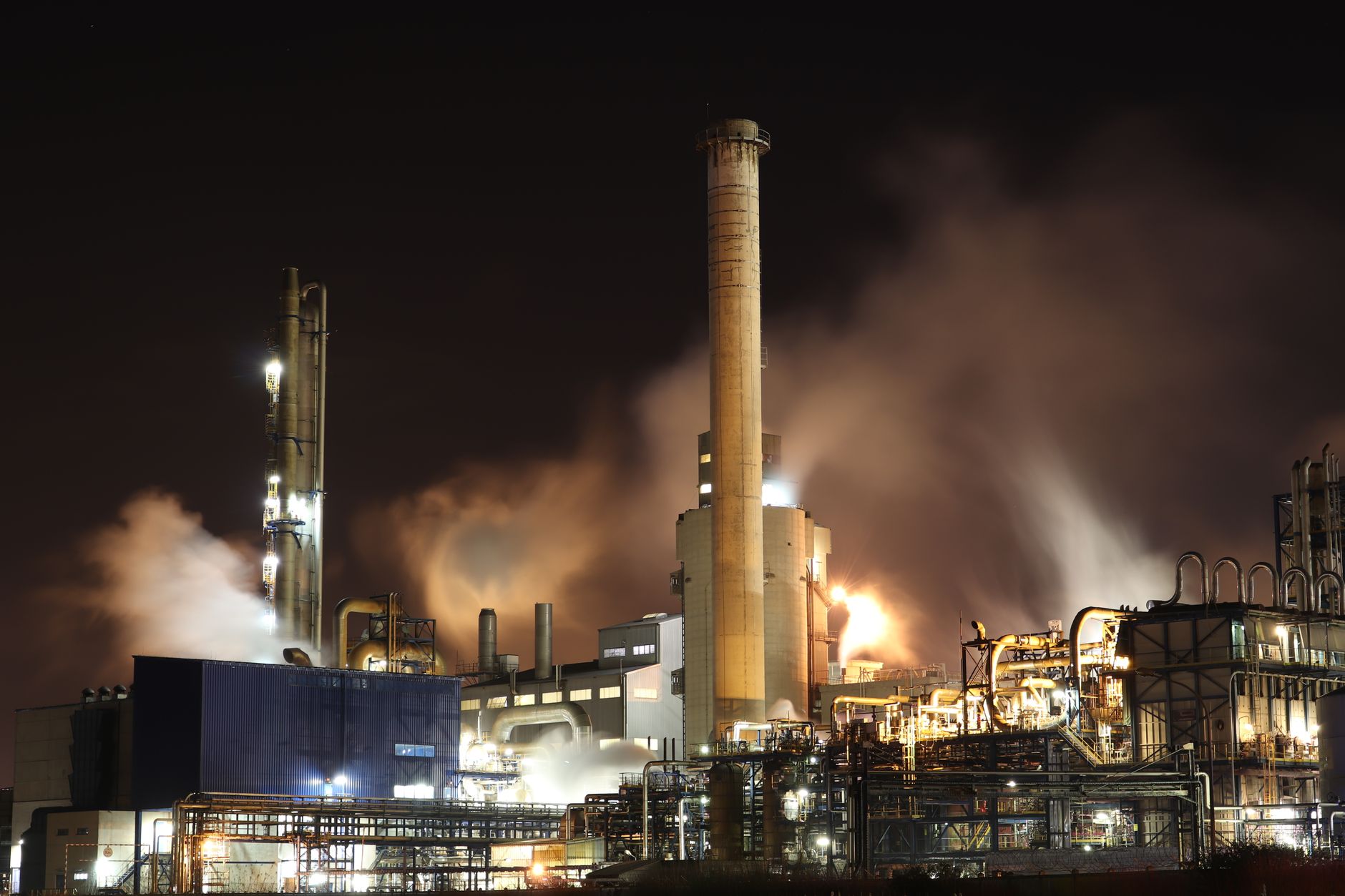 aerial view of industrial oil processing towers and pipelines in a desert landscape with plumes of dark smoke visible on the horizon