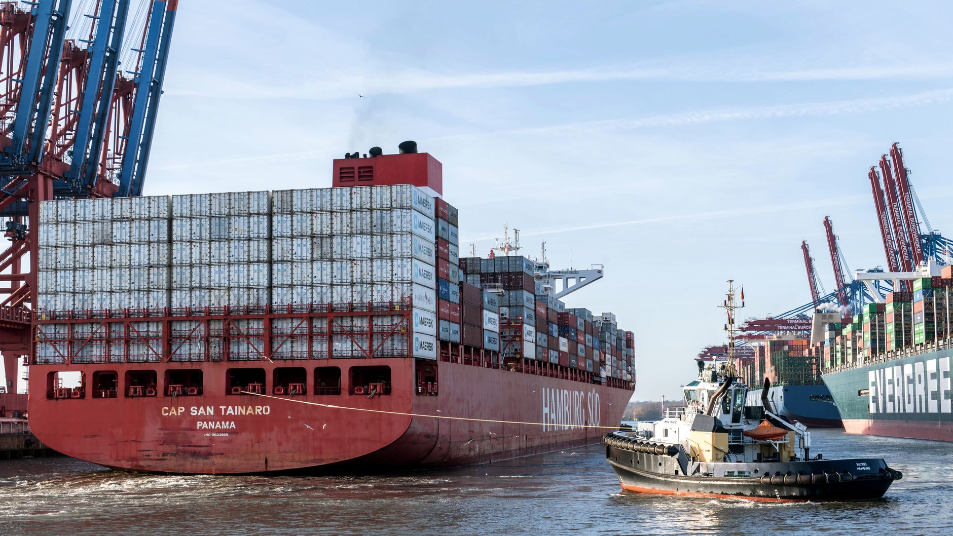 container ship navigating through a narrow strait with military vessels visible in the background against a grey sky