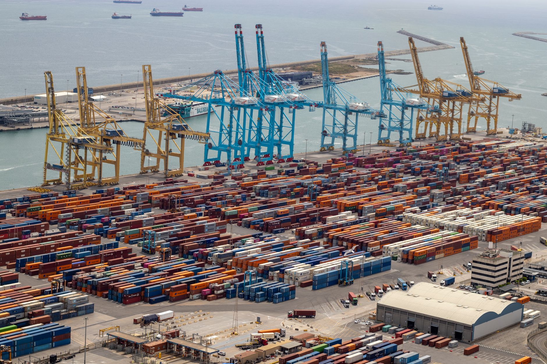 exterior view of a commercial port with container cranes and stacked cargo units along a Mediterranean quayside at midday