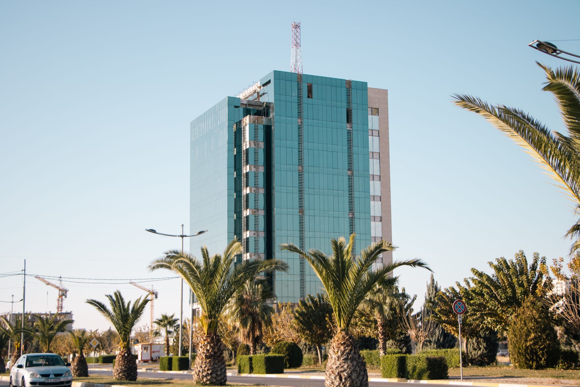 exterior of a modern glass-fronted technology campus in Tel Aviv's HaKirya district at midday, with palm trees and security barriers visible in the foreground