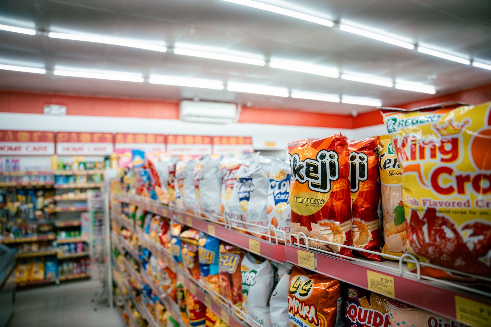wide interior shot of a large Japanese superstore aisle lined with packaged food products and price label signage, brightly lit with empty shopping carts visible in the foreground