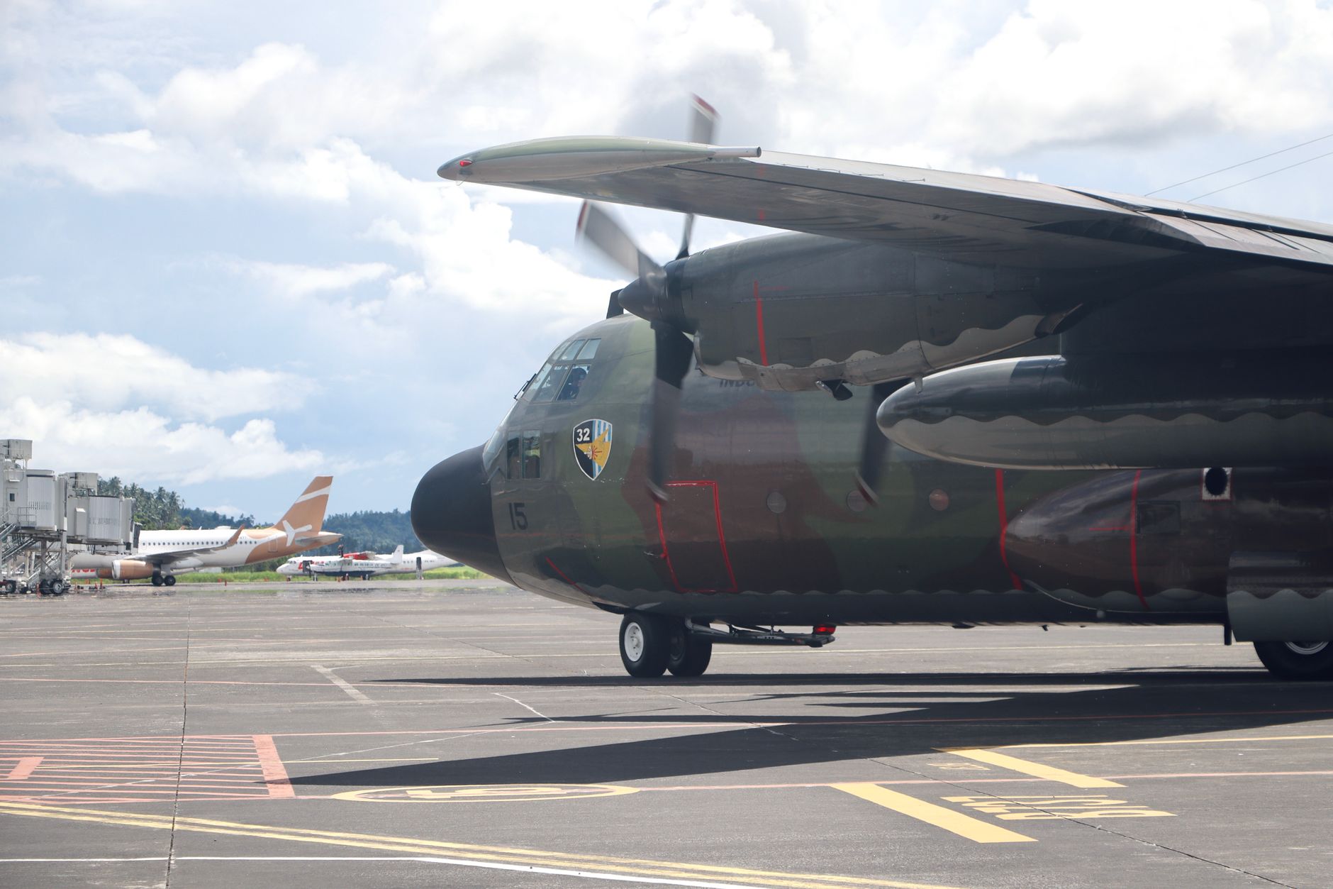a precision-guided munition mounted beneath the wing of a military aircraft on a sun-lit airfield tarmac, ground crew equipment visible in the background
