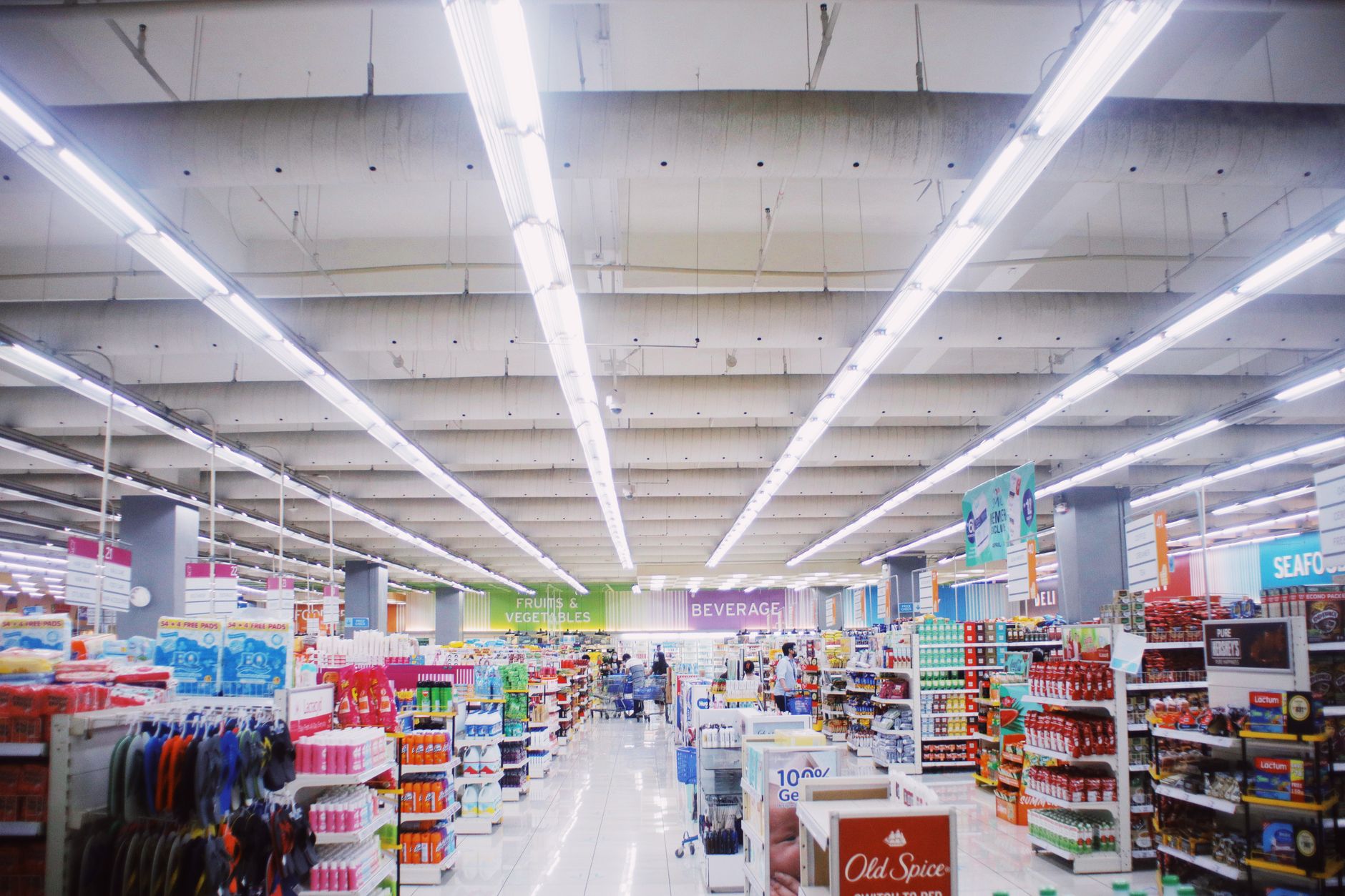 a grocery store aisle with price tags clearly visible on shelves stocked with everyday food items, fluorescent lighting overhead, shopping carts lined up near the entrance