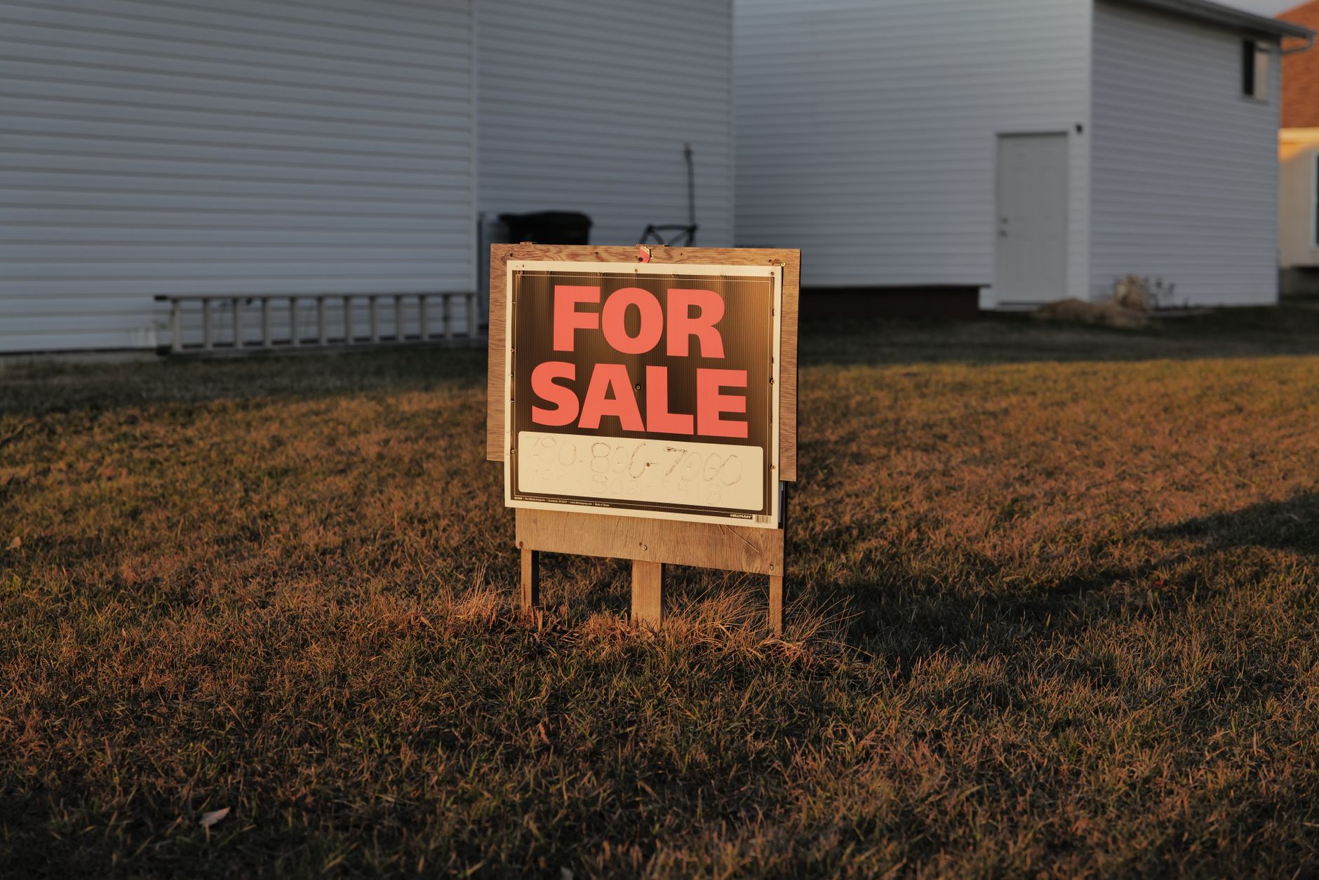 a row of residential homes on a suburban street with a real estate for-sale sign in the foreground, late afternoon light casting long shadows across the lawn
