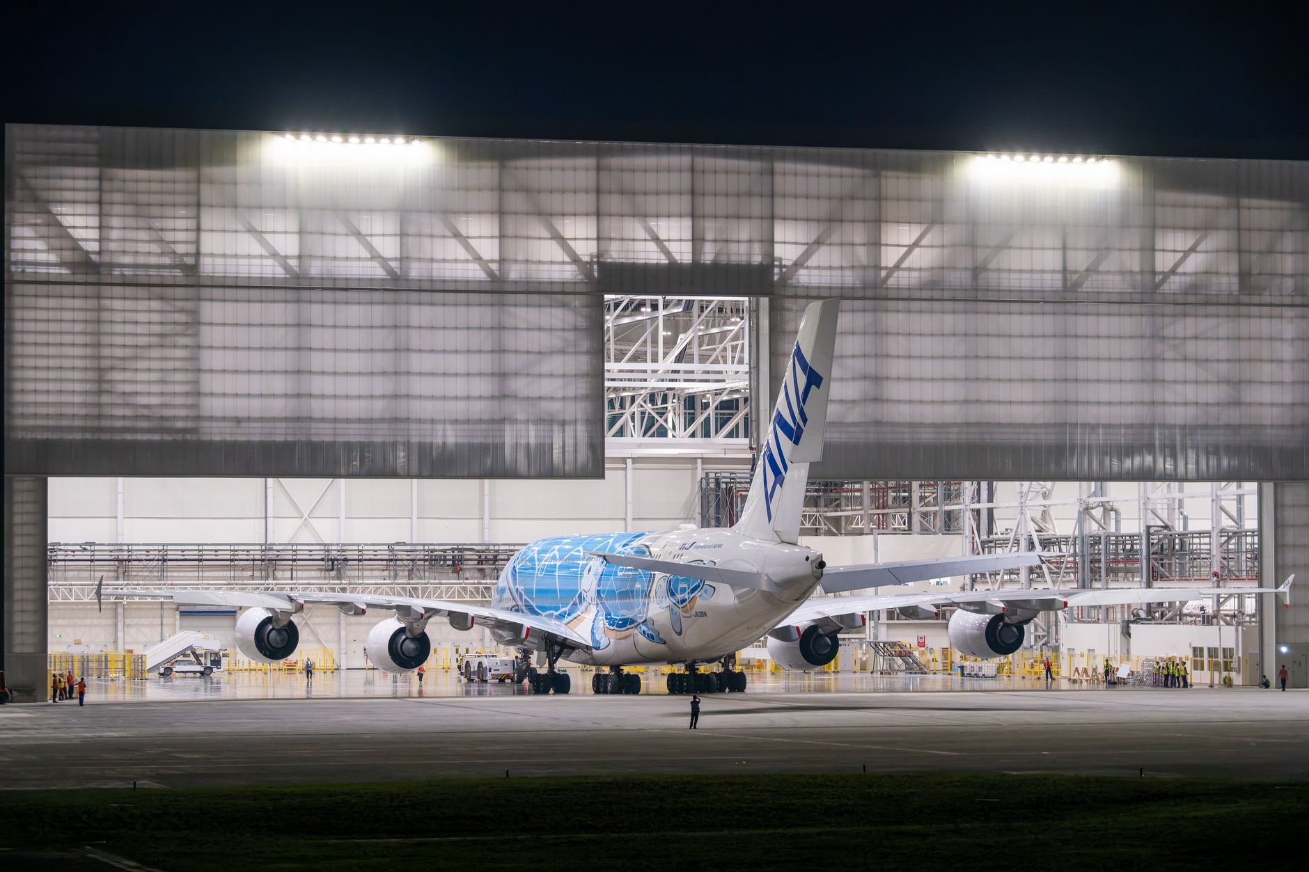 exterior of a large aerospace manufacturing facility with aircraft components visible through an open hangar door at midday