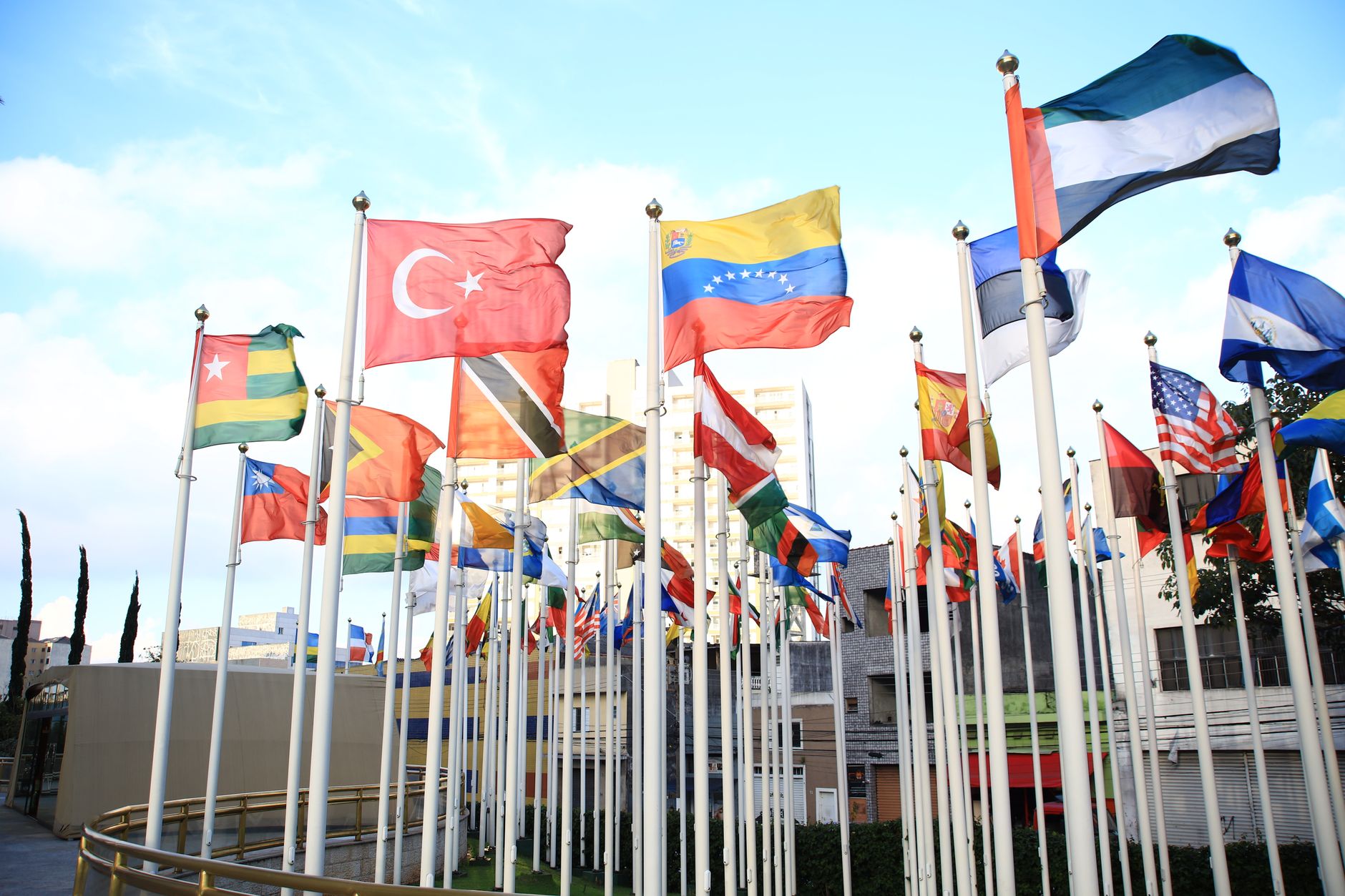 wide-angle exterior view of an international conference centre with national flags lining the entrance forecourt under overcast skies