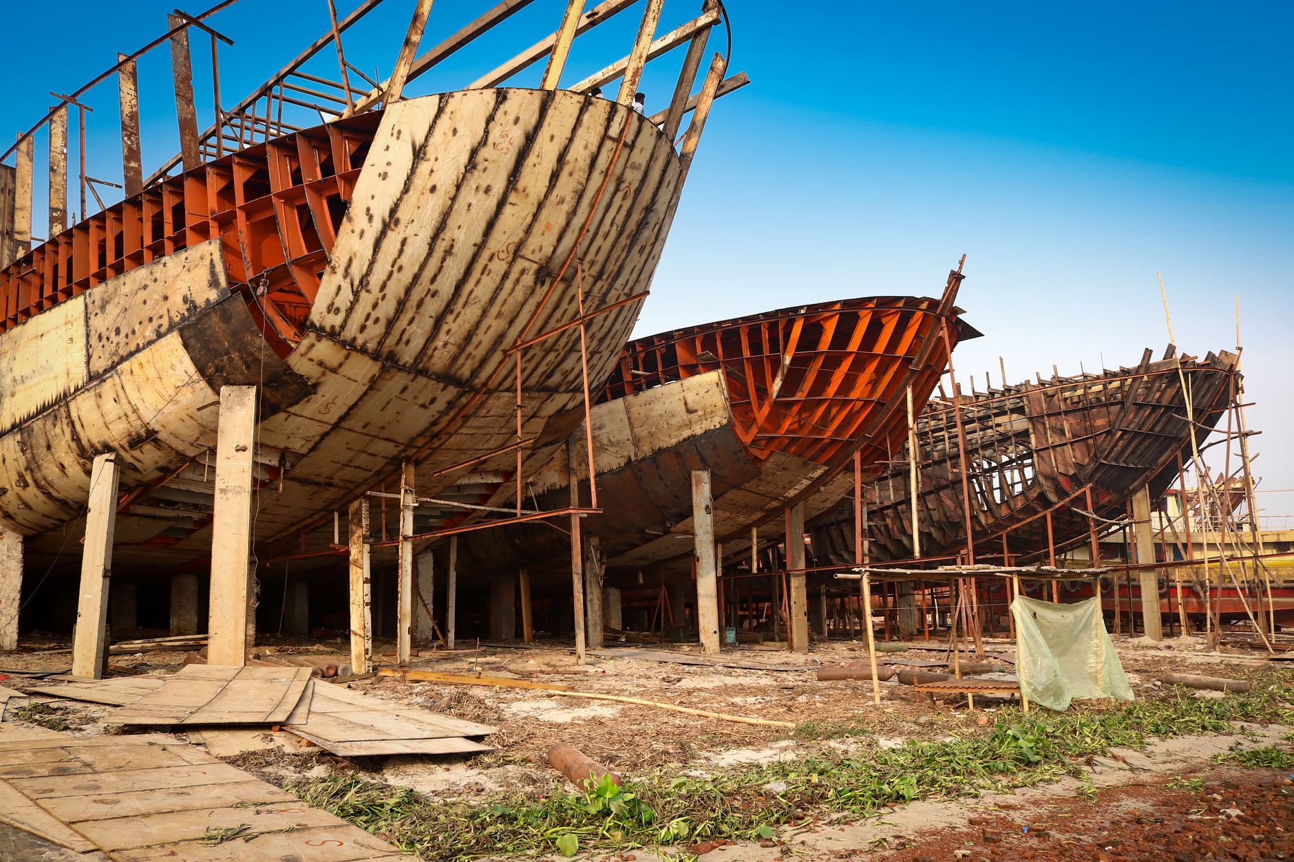 wide-angle view of a large naval shipyard at dusk, dry docks illuminated by industrial floodlights with the skeletal hull of a destroyer under construction visible in the foreground