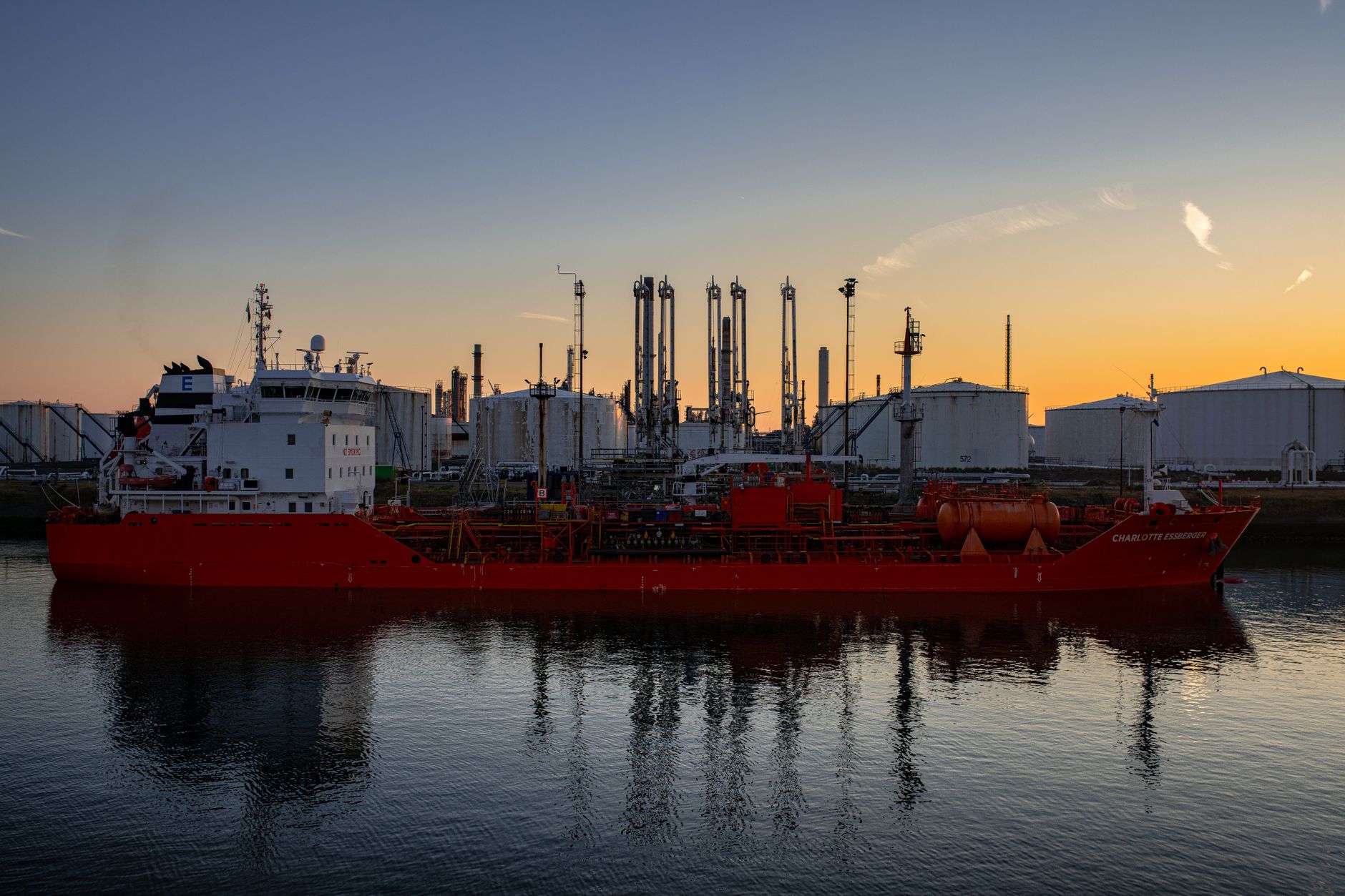 exterior of a large oil refinery complex at dusk, with flare stacks illuminated against an orange sky and tanker berths visible in the distance