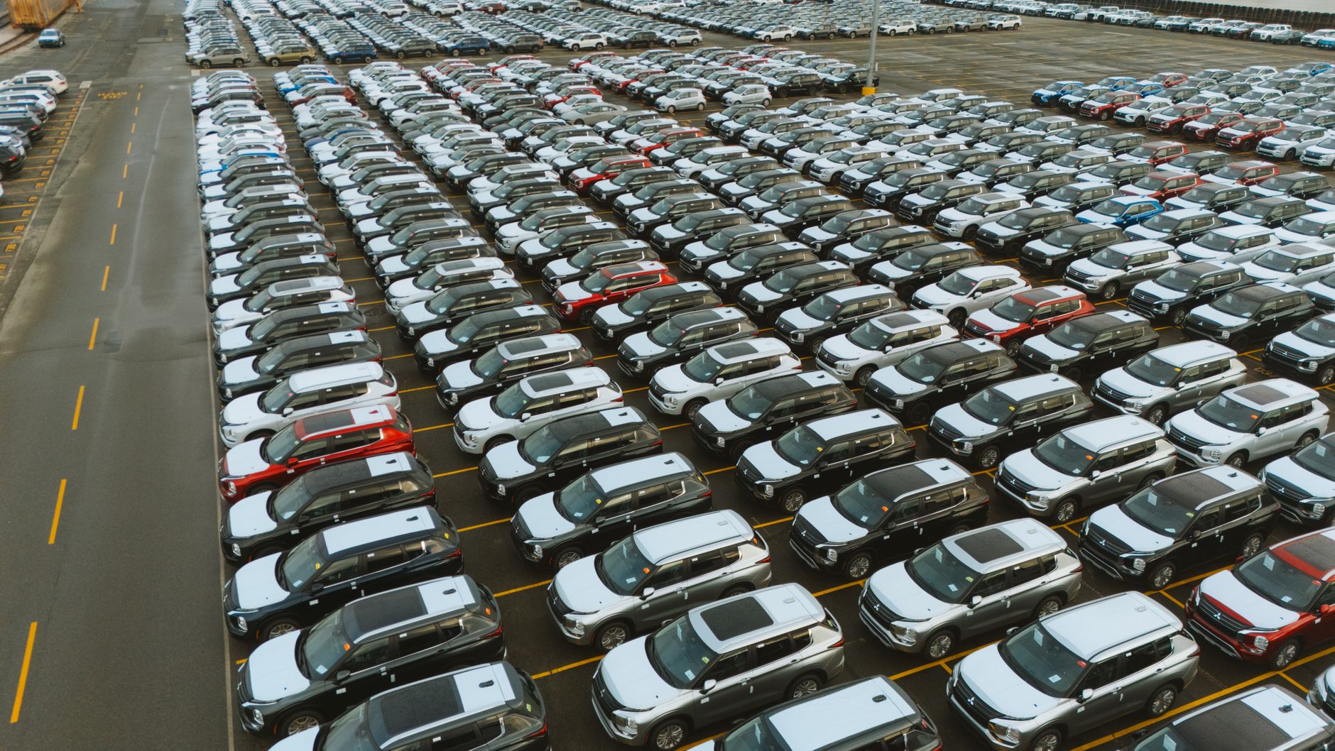 rows of electric vehicles on an outdoor delivery lot at a European port terminal, cargo ships visible in the background